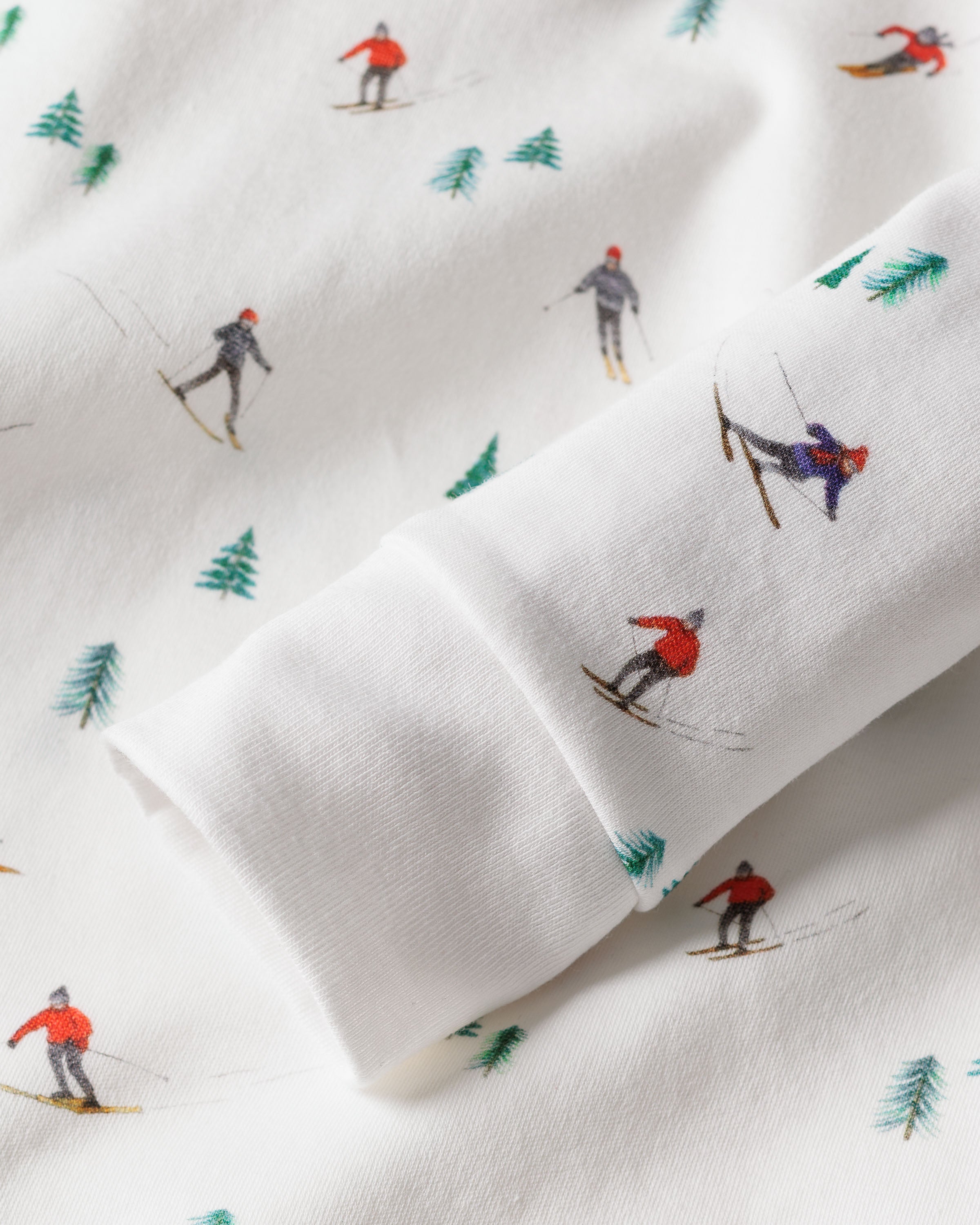 A close-up of Petite Plumes Après Ski Kids Pima Snug Fit Pajama Set showcases skiers in vibrant attire, moving in various directions on soft and cozy white fabric, dotted with small green pine trees—ideal for a snug long sleeve top or pant set.
