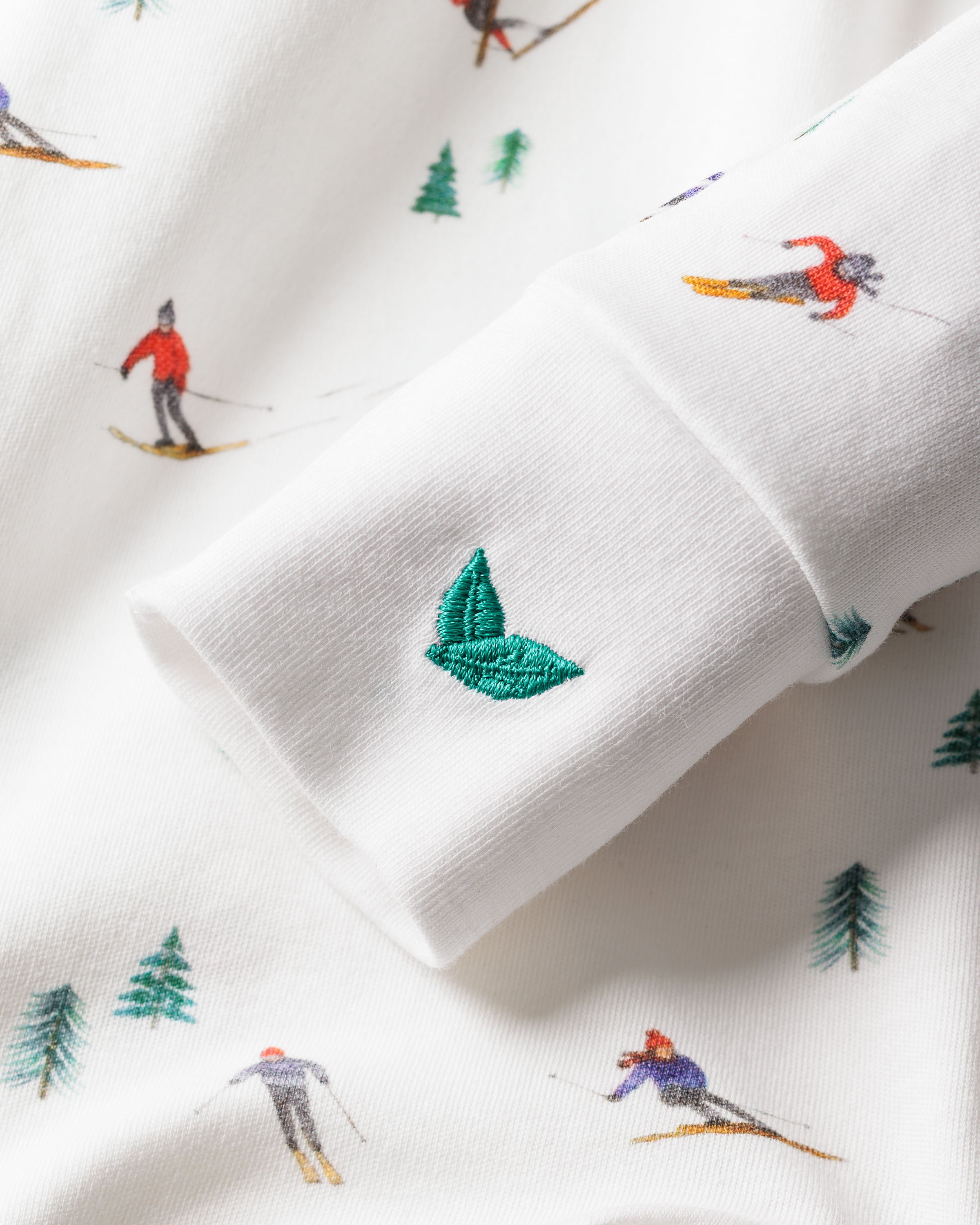Close-up of Petite Plumes Babys Pima Romper in Après Ski, showcasing a small embroidered green leaf on soft white Pima cotton fabric. The pattern includes skiers in vibrant red and blue attire with green pine trees. Leaf emblem is the focus.