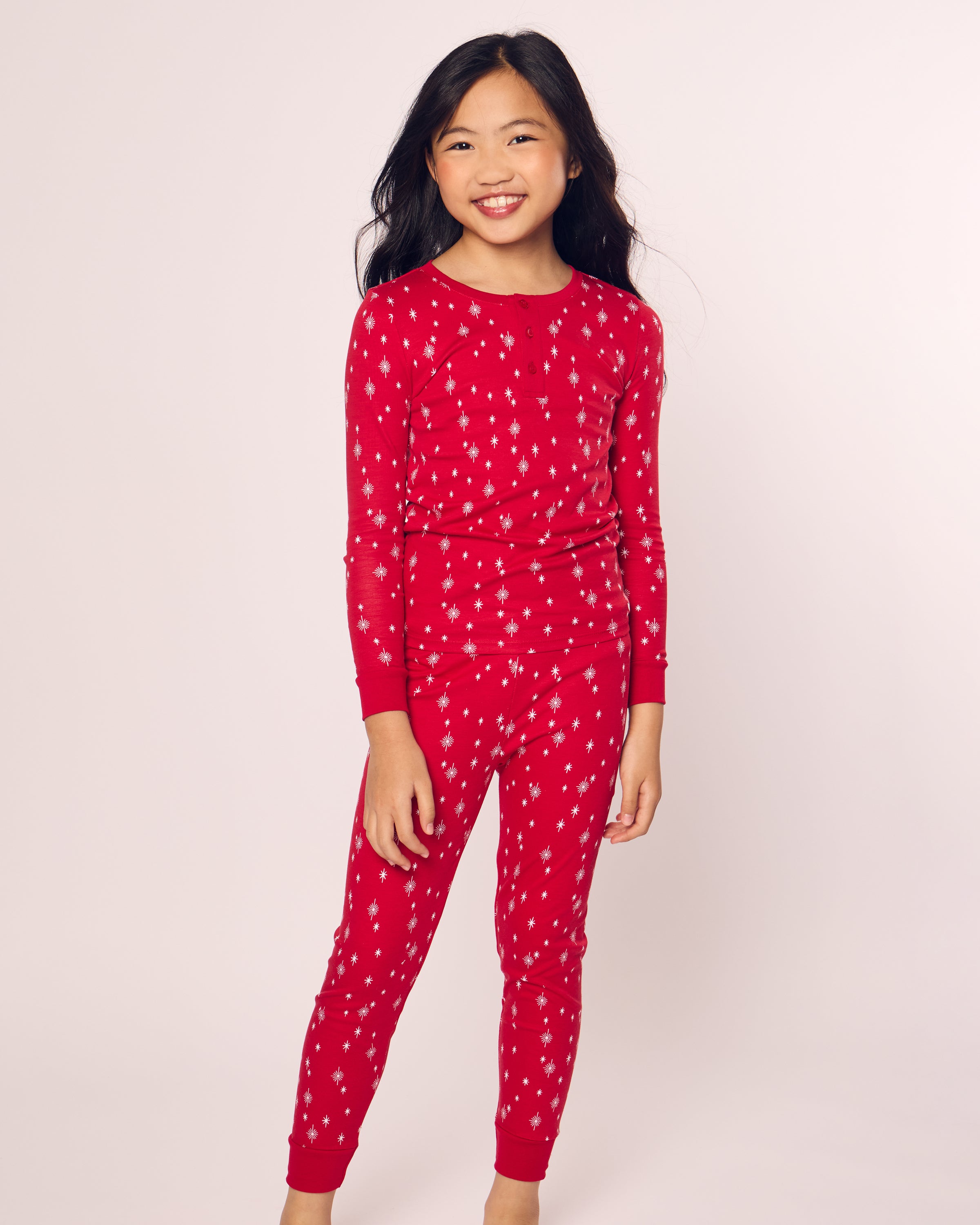 A young girl smiles against a plain background, wearing the Kids Pima Snug Fit Pajama Set in Starry Night by Petite Plume. Her long dark hair cascades down, and the cozy red pajamas with white star patterns include a comfy long-sleeve top made of soft pima cotton, wrapping her in warmth.