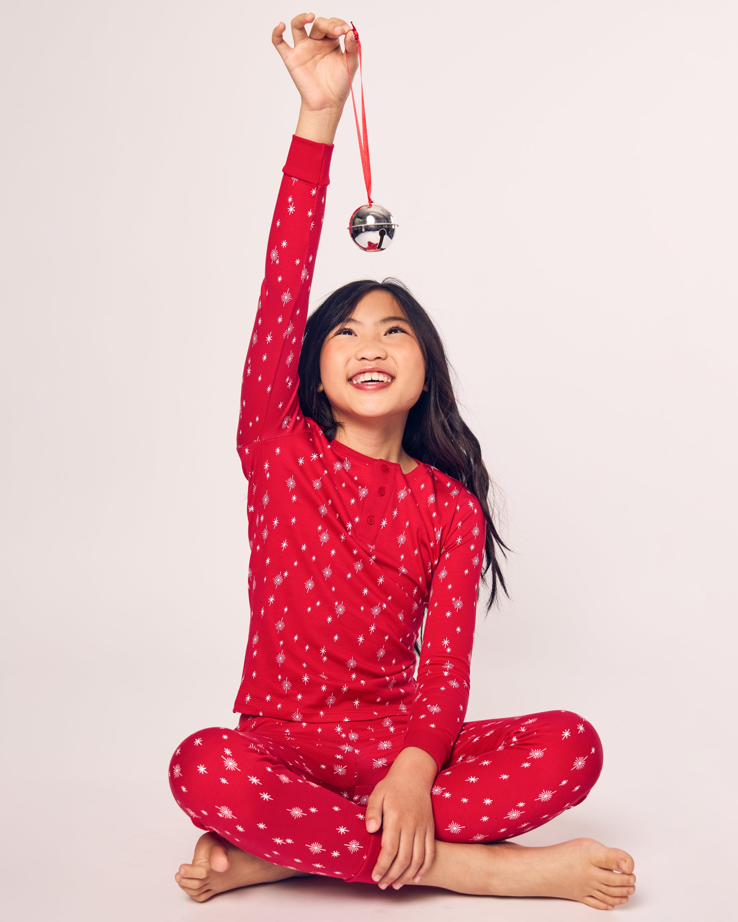A child, wearing Petite Plumes Kids Pima Snug Fit Pajama Set in Starry Night, sits cross-legged and smiles while holding a silver jingle bell by a red ribbon against a plain white background.