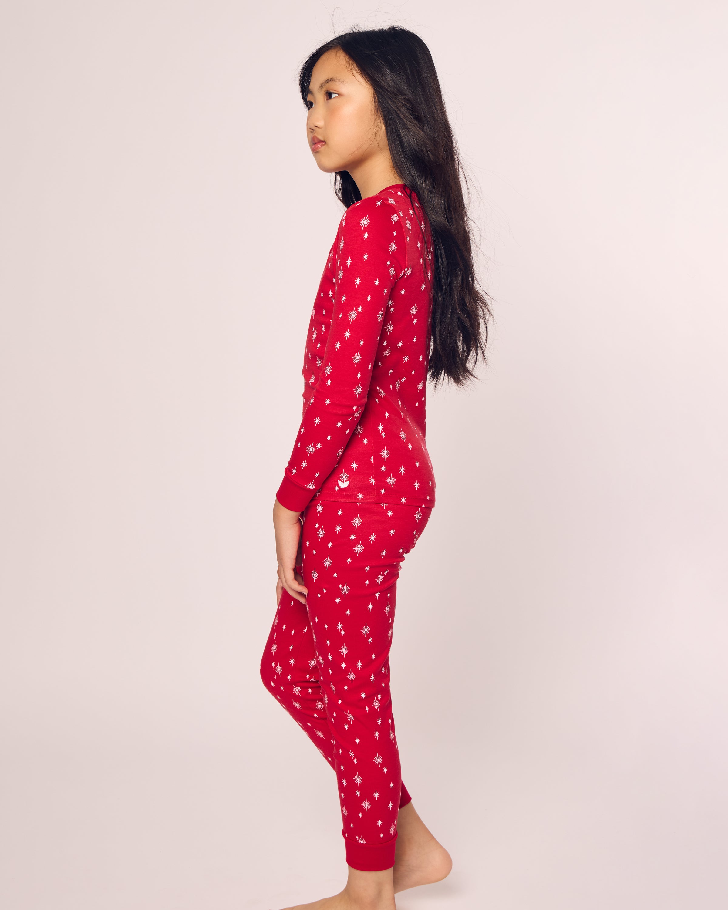 A young girl with long dark hair stands in profile, wearing Petite Plumes Kids Pima Snug Fit Pajama Set in Starry Night, featuring a cozy red design with white snowflakes. The soft Pima cotton long-sleeve top complements her barefoot pose against a plain off-white background.