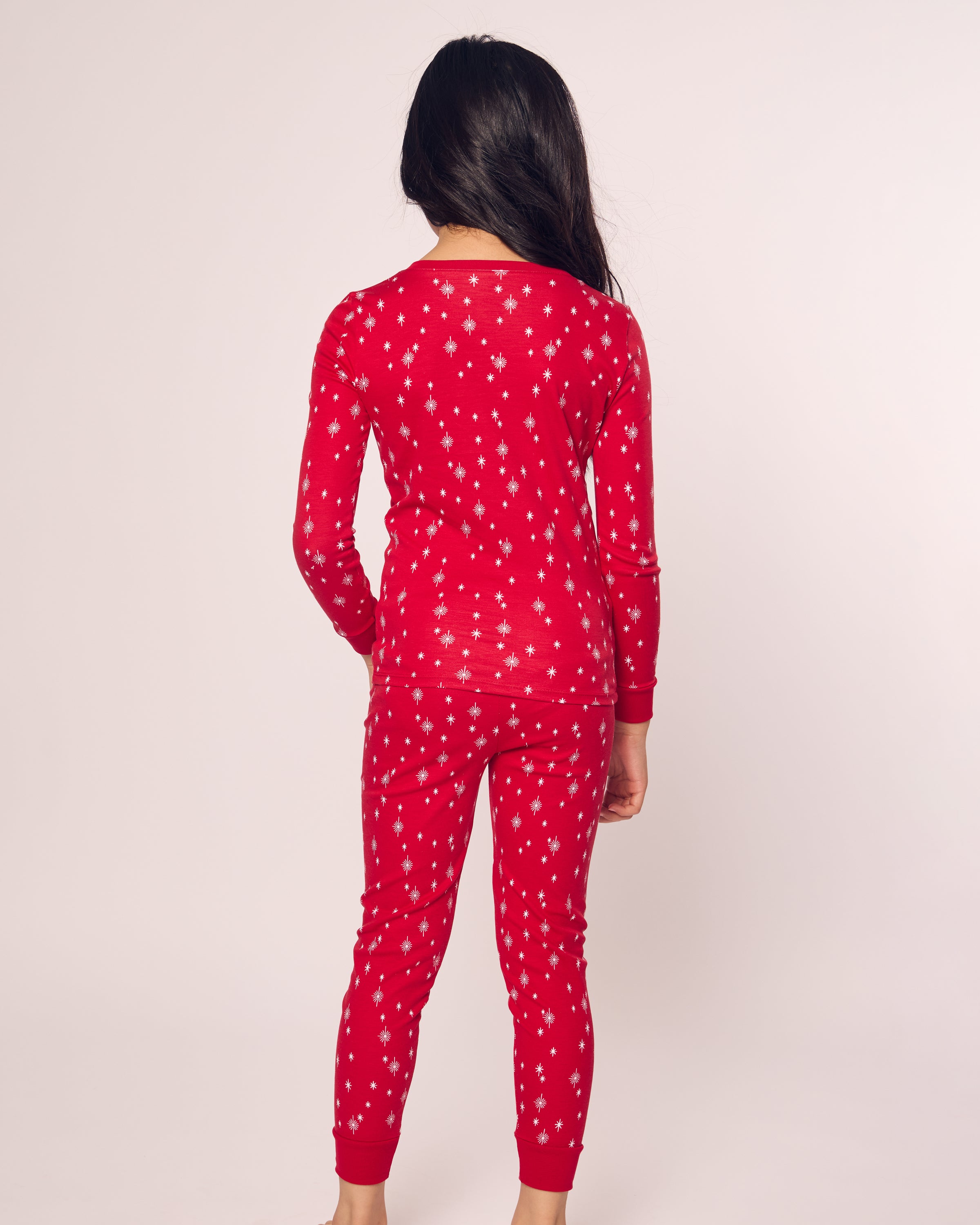 A person with long dark hair stands facing away, wearing a long sleeve top and red pajamas featuring a white snowflake pattern. Crafted from soft Pima cotton by Petite Plume, their Kids Pima Snug Fit Pajama Set in Starry Night exudes comfort against a plain, light-colored background.