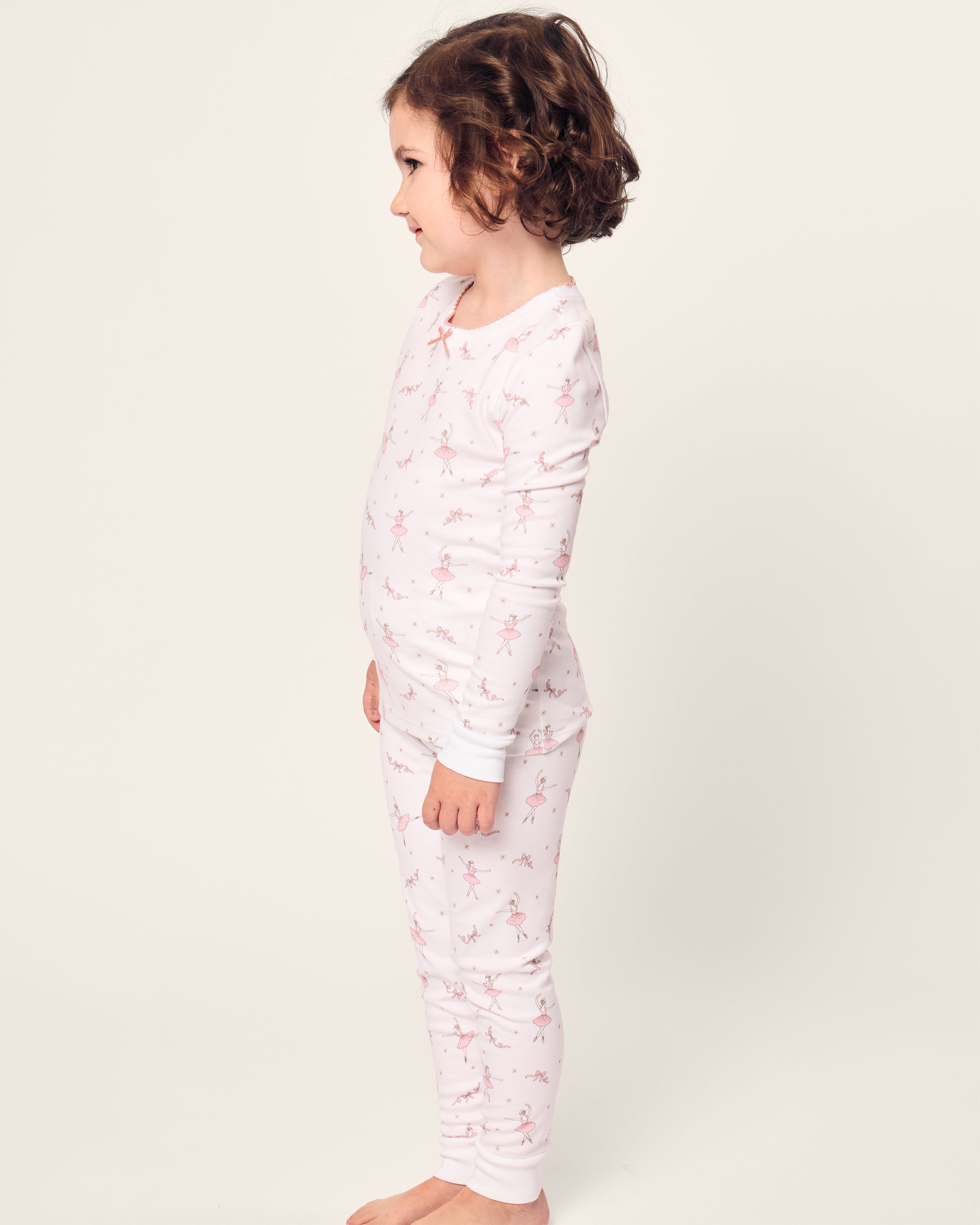 A child with curly hair, wearing Petite Plumes Kids Pima Snug Fit Pajama Set in Sugar Plum Fairy, stands in profile. The soft white pajamas feature a pink floral pattern and cozy long sleeves with matching pants, set against a soft neutral background that highlights the childs stance.