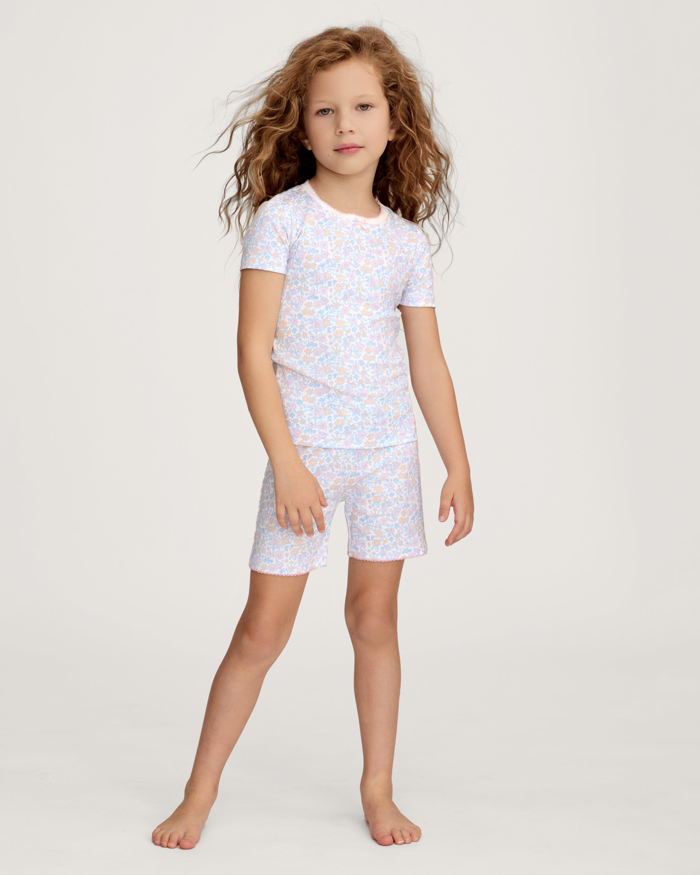 A young girl with long, wavy hair smiles with her hands on her hips, wearing the Kid's Pima Snug Fit Short Set in Fleur Blanche. She stands against a plain, light-colored background.
