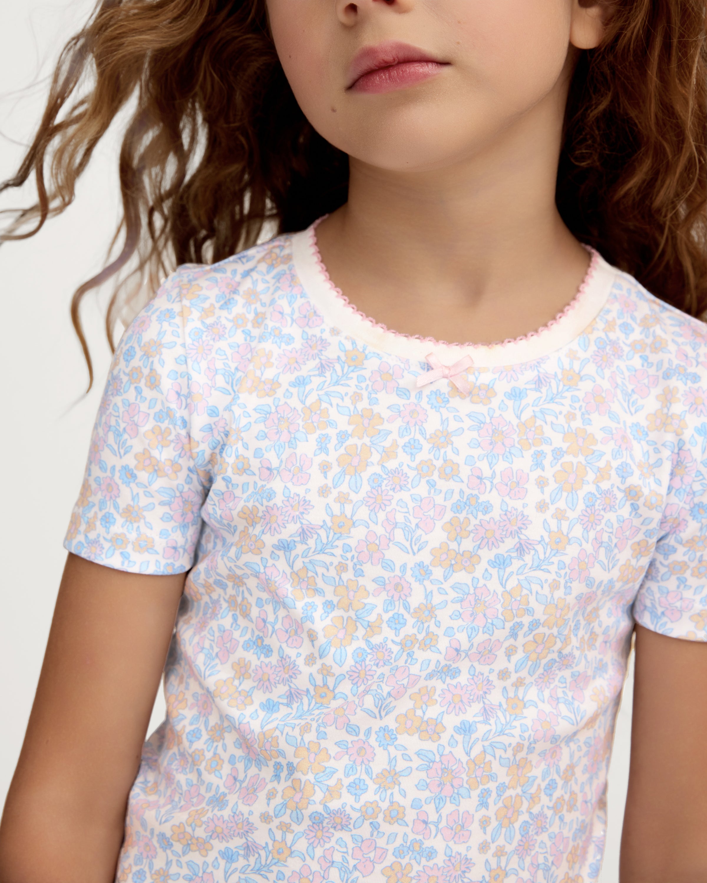 A young girl with long, wavy hair smiles with her hands on her hips, wearing the Kid's Pima Snug Fit Short Set in Fleur Blanche. She stands against a plain, light-colored background.