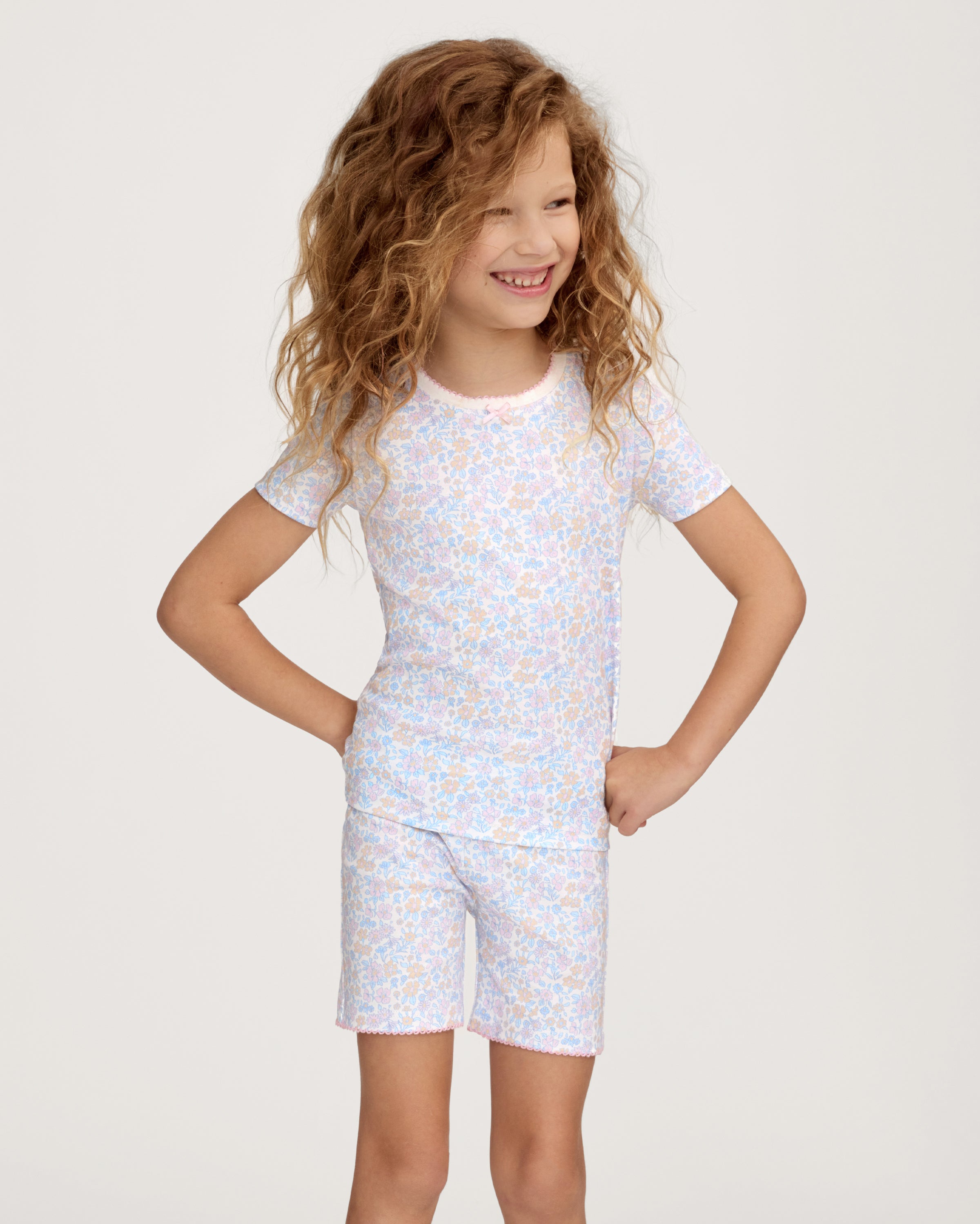 A young girl with long, wavy hair smiles with her hands on her hips, wearing the Kid's Pima Snug Fit Short Set in Fleur Blanche. She stands against a plain, light-colored background.