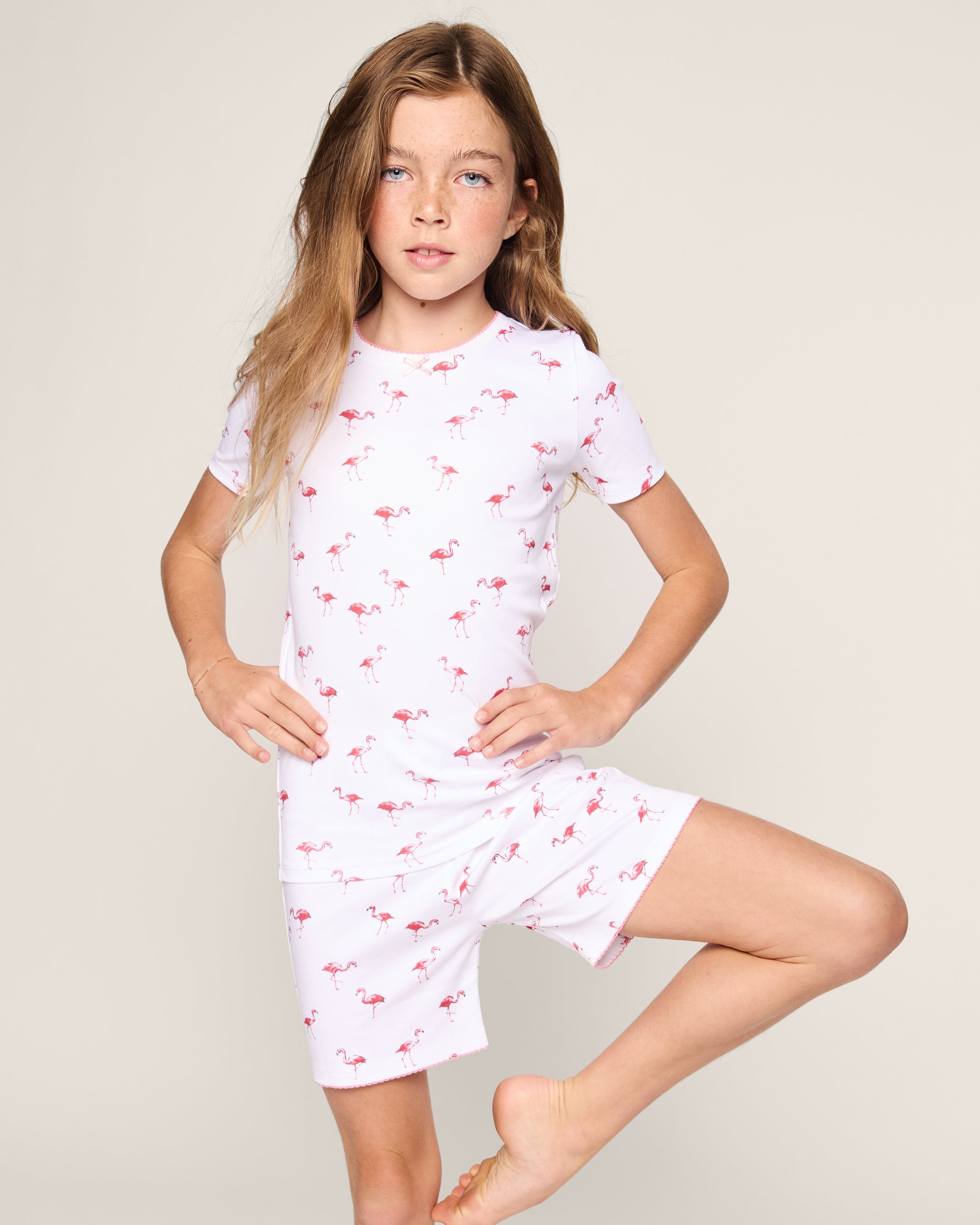 A young girl with long hair stands on one leg, wearing the Petite Plume Kids Pima Snug Fit Short Set in Flamingos, against a plain light background.