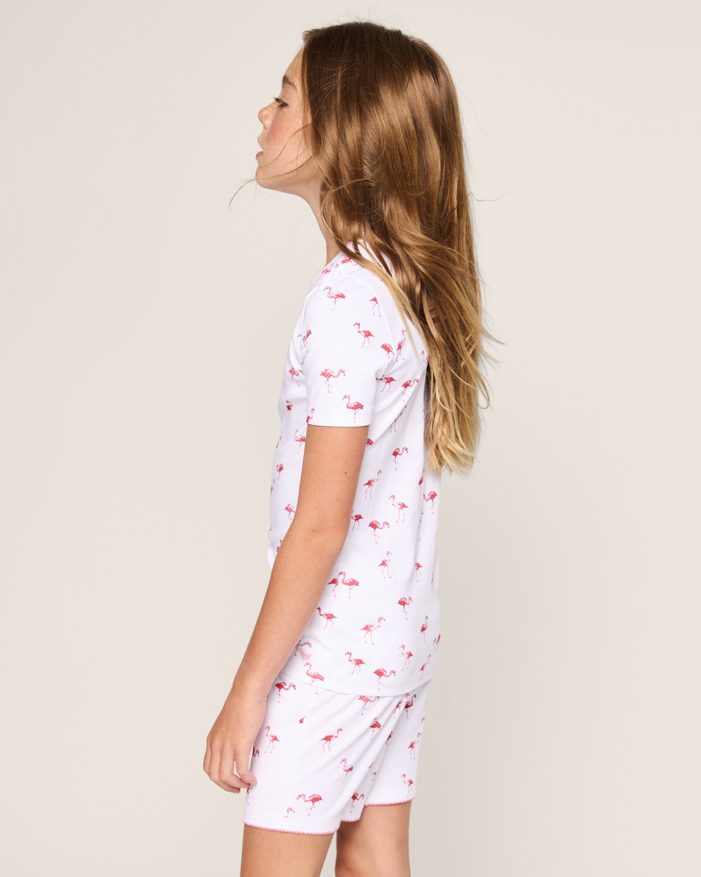 A young girl with long brown hair stands in profile, wearing the Petite Plume Kids Pima Snug Fit Short Set in Flamingos—white pajamas with a pink flamingo print—against a plain light background.