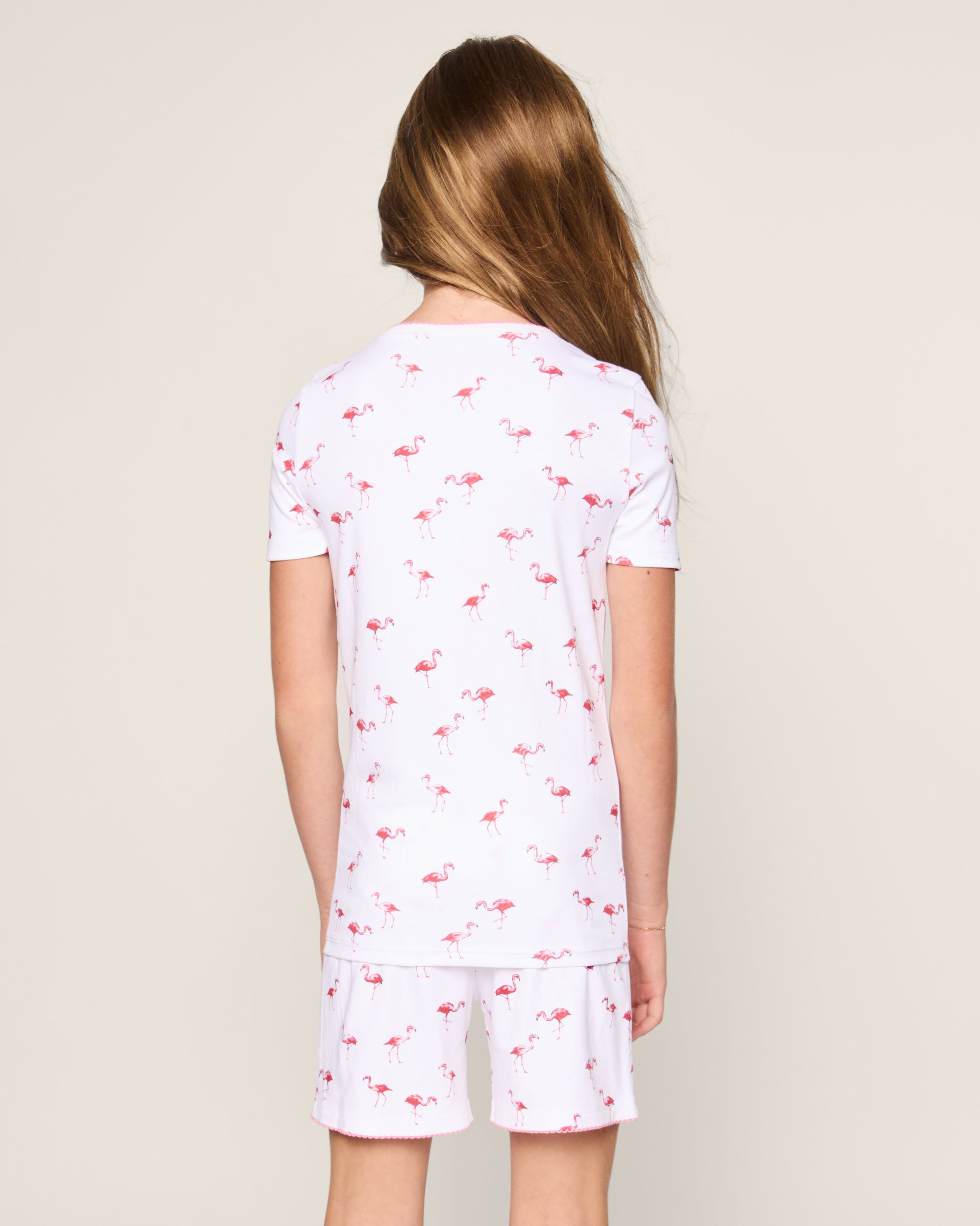 A young girl with long brown hair stands facing away, wearing the Petite Plume Kids Pima Snug Fit Short Set in Flamingos—white pajamas with a pink flamingo print—crafted from soft Pima cotton, set against a plain beige background.