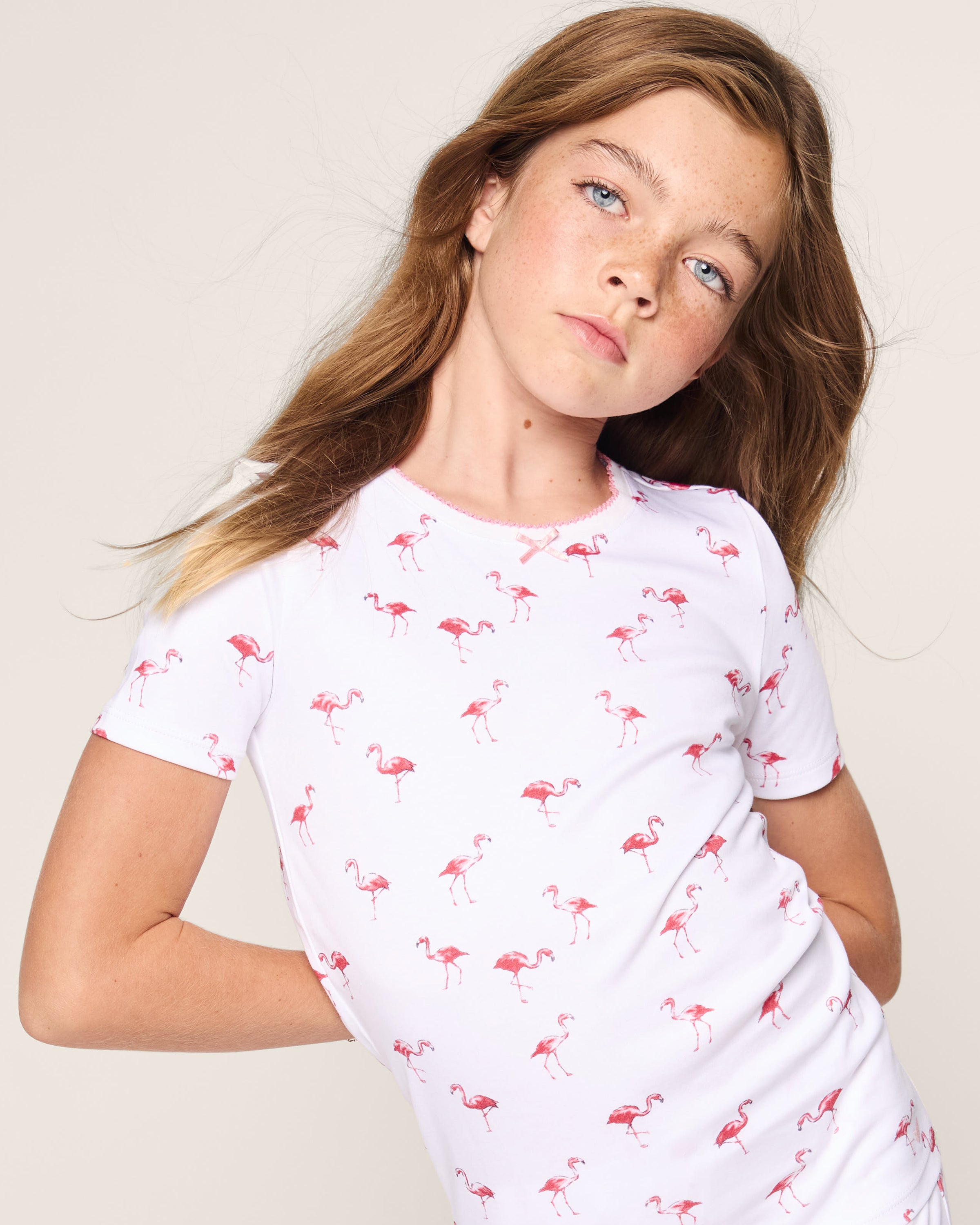 A young girl wears the Petite Plume Kids Pima Snug Fit Short Set in Flamingos, featuring a white background with pink flamingo patterns, standing against a plain light background.