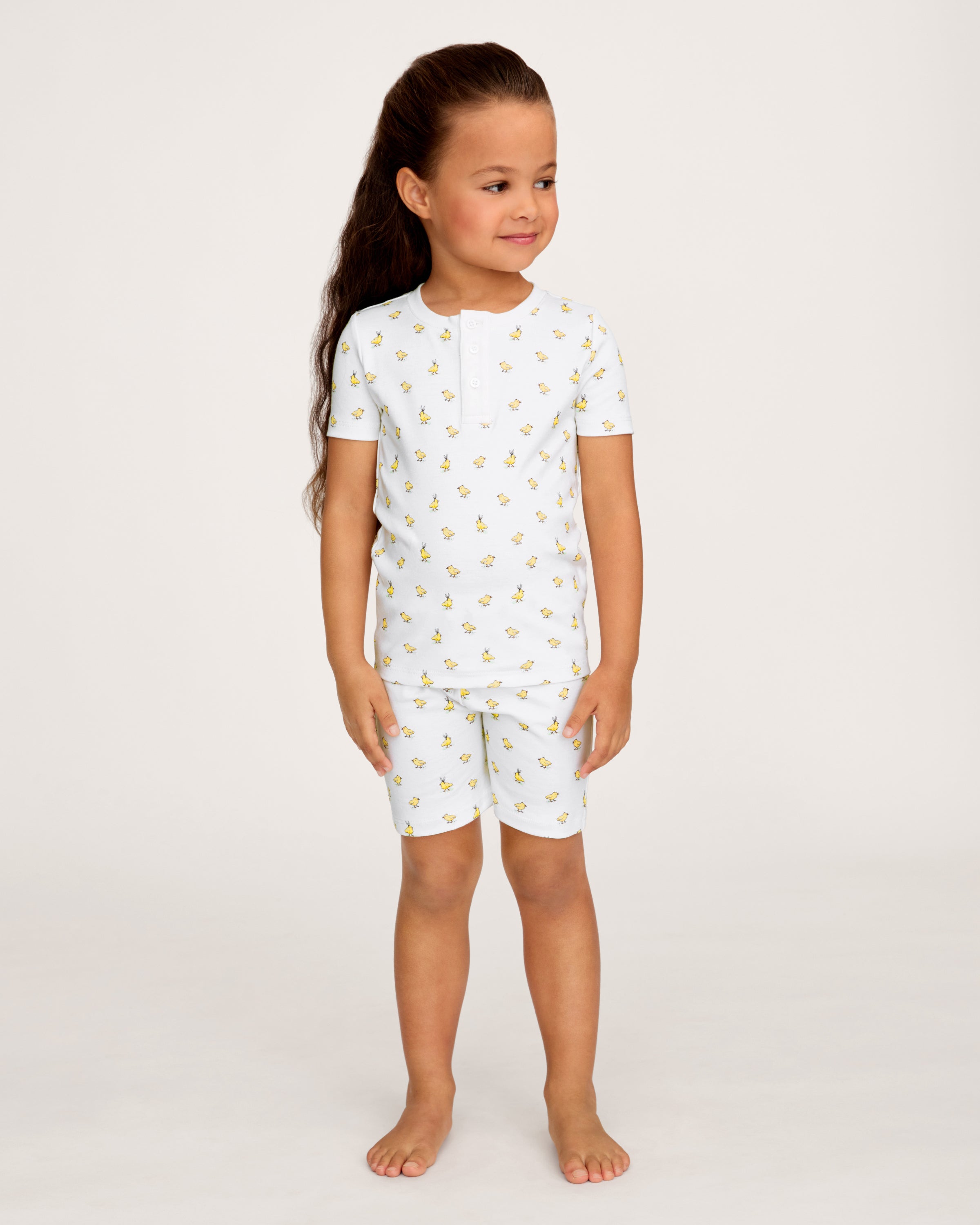 A young child with long brown hair smiles while wearing the Kid's Pima Snug Fit Short Set in Très Chick, standing against a plain light background.