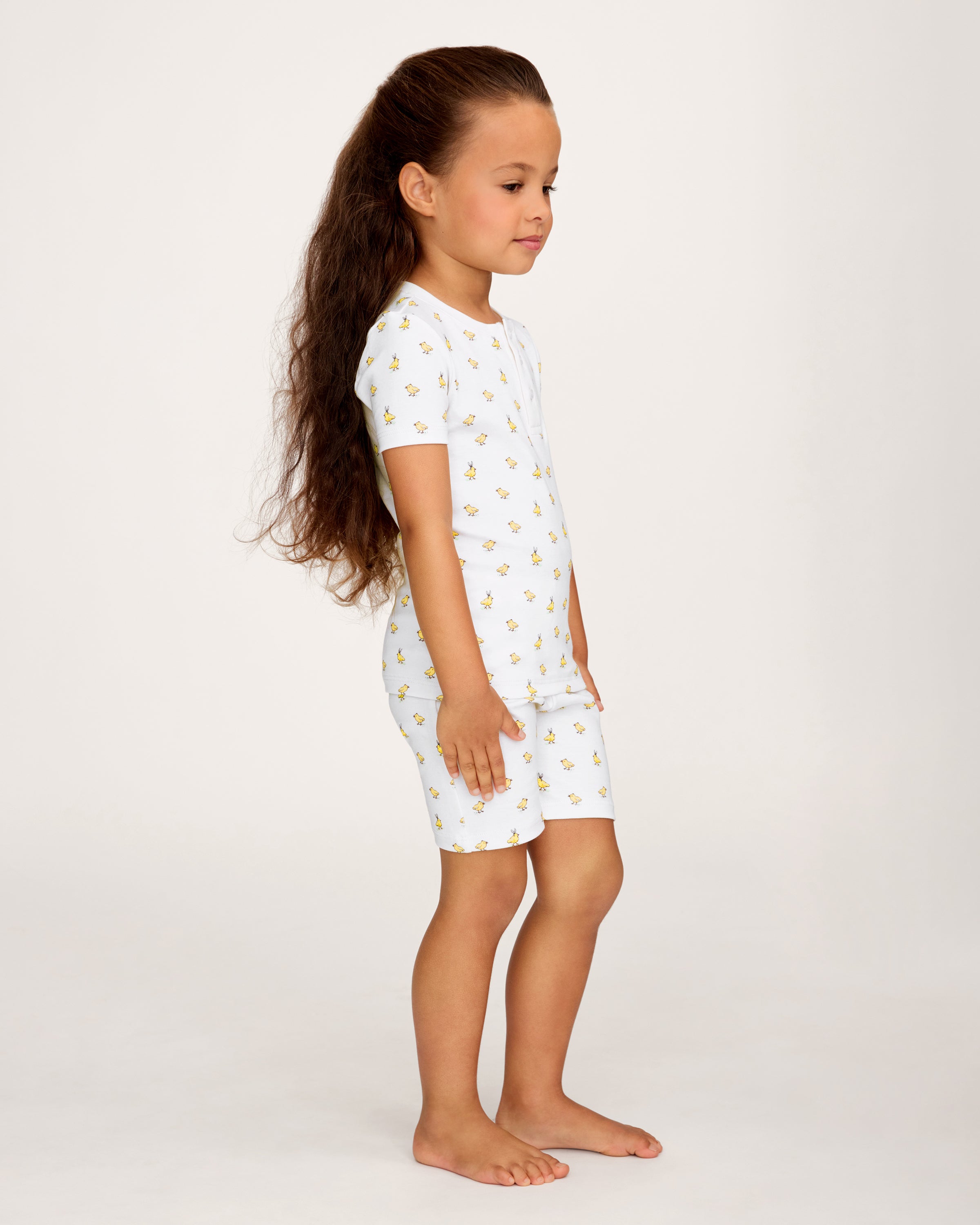 A young child with long brown hair smiles while wearing the Kid's Pima Snug Fit Short Set in Très Chick, standing against a plain light background.
