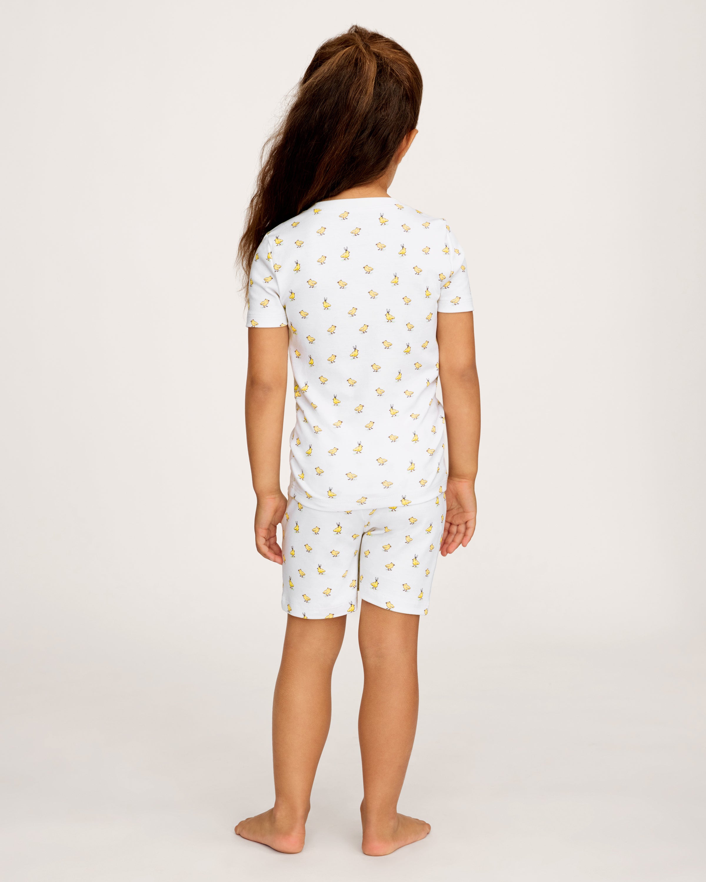 A young child with long brown hair smiles while wearing the Kid's Pima Snug Fit Short Set in Très Chick, standing against a plain light background.
