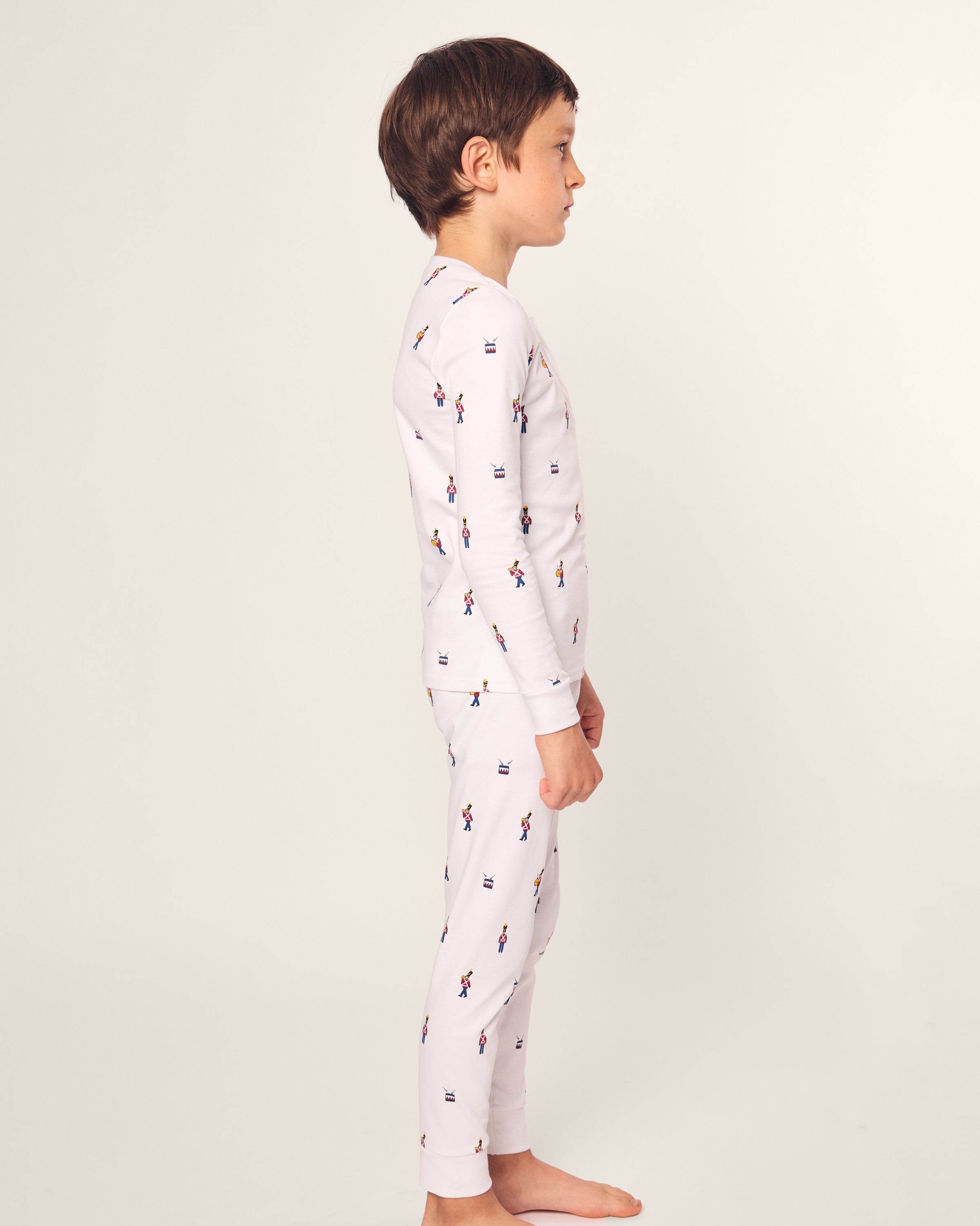 A child with short brown hair, barefoot on an off-white background, models the luxurious Petite Plume Kids Pima Snug Fit Pajama Set in Toy Soldier. Crafted from soft Pima Cotton, this matching white set features a long sleeve top and pants adorned with multicolored skater illustrations.