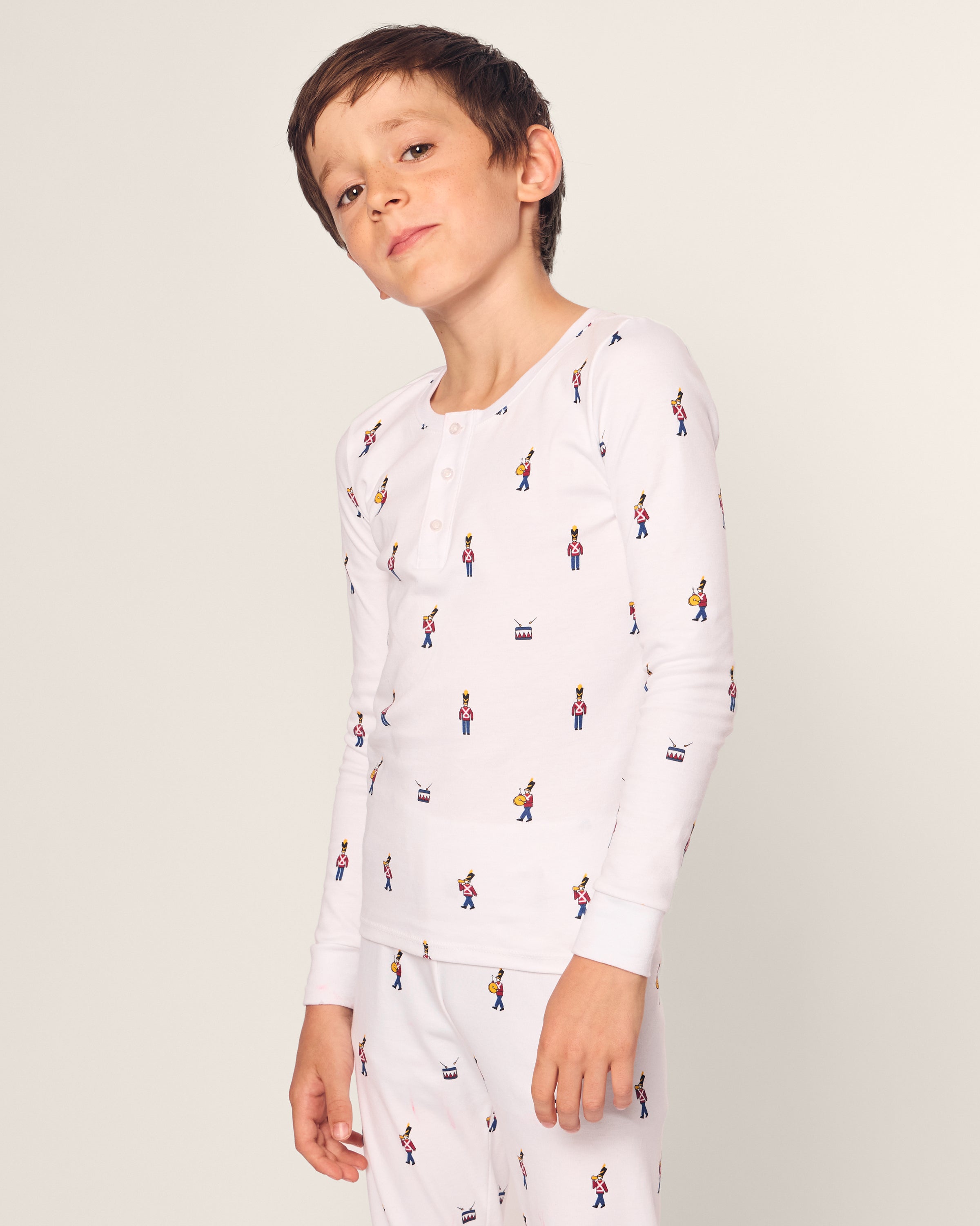 A young boy wearing Petite Plumes Kids Pima Snug Fit Pajama Set in Toy Soldier, made from luxurious Pima Cotton, stands with a slight smile. The white pajamas have cartoon character prints and a button-up design, set against a plain, light-colored background.
