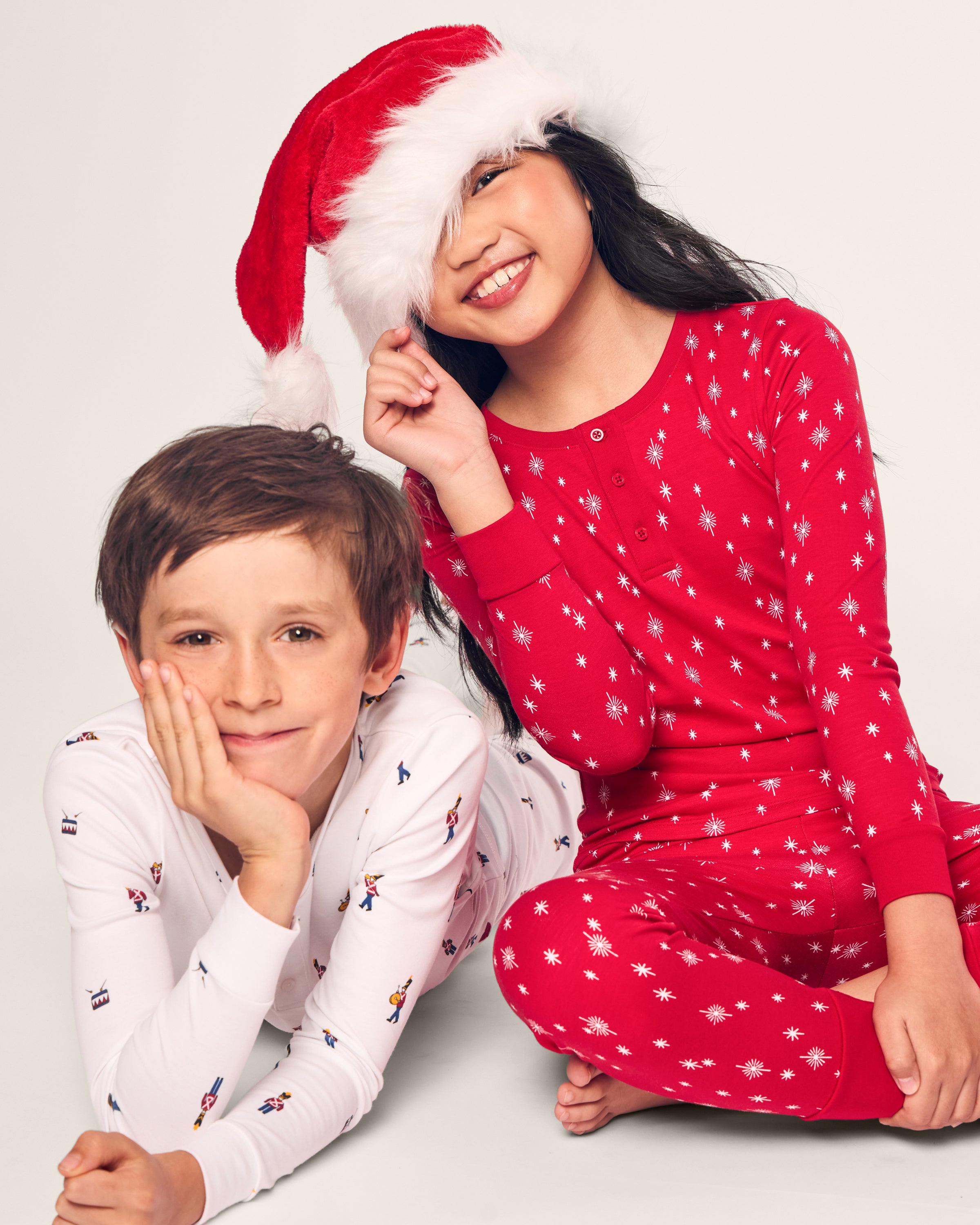 Two children wearing luxurious sleepwear sit on the floor. One smiles playfully in a red long sleeve top and pant set with a Santa hat, while the other looks content in Petite Plumes Kids Pima Snug Fit Pajama Set in Toy Soldier. The background is plain.
