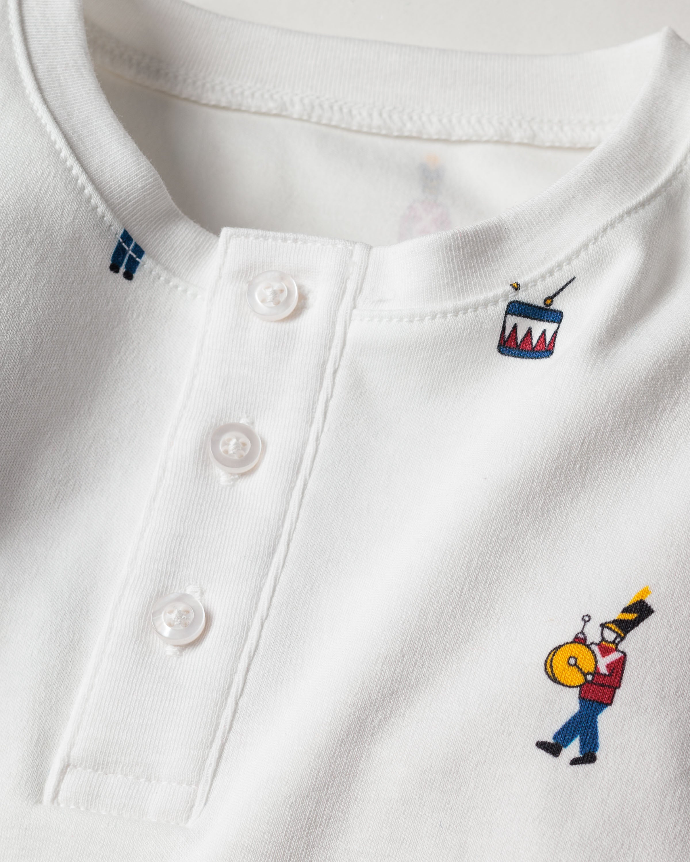Close-up of a Petite Plume Kids Pima Snug Fit Pajama Set in Toy Soldier. Features small prints on luxurious white Pima cotton, with colorful drum by the neckline and soldier with a yellow drum at the bottom. Includes three white buttons on the placket, perfect for pairing with matching set.