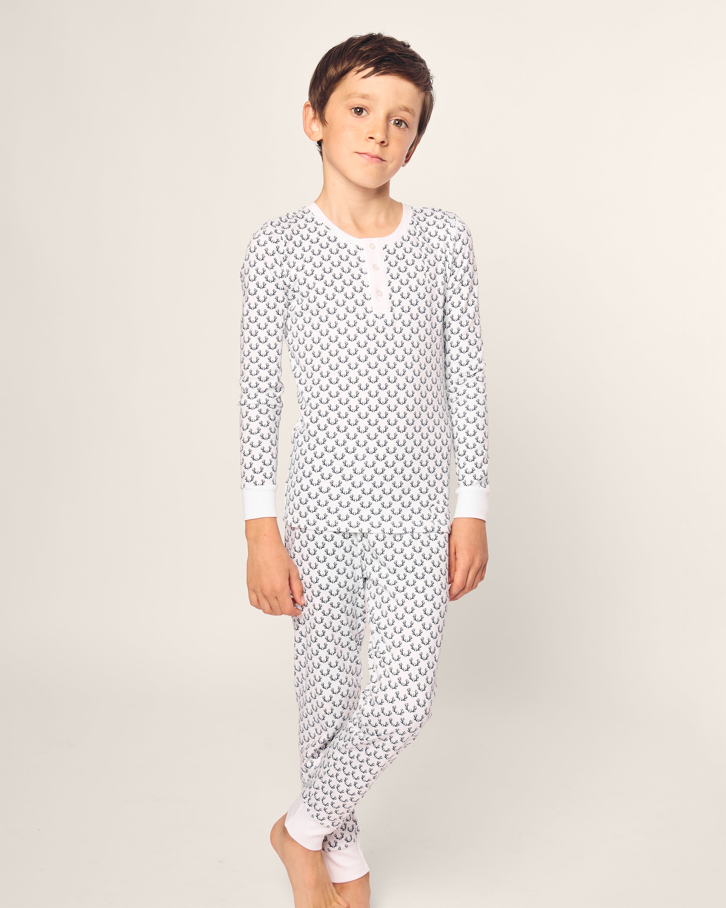 A child models Petite Plumes Kids Pima Snug Fit Pajama Set in Nordic Antlers on white, featuring long sleeves and chest buttons. The soft Pima cotton onesie displays black crescent designs against a light background, as the barefoot child, with a neutral expression, faces the camera.