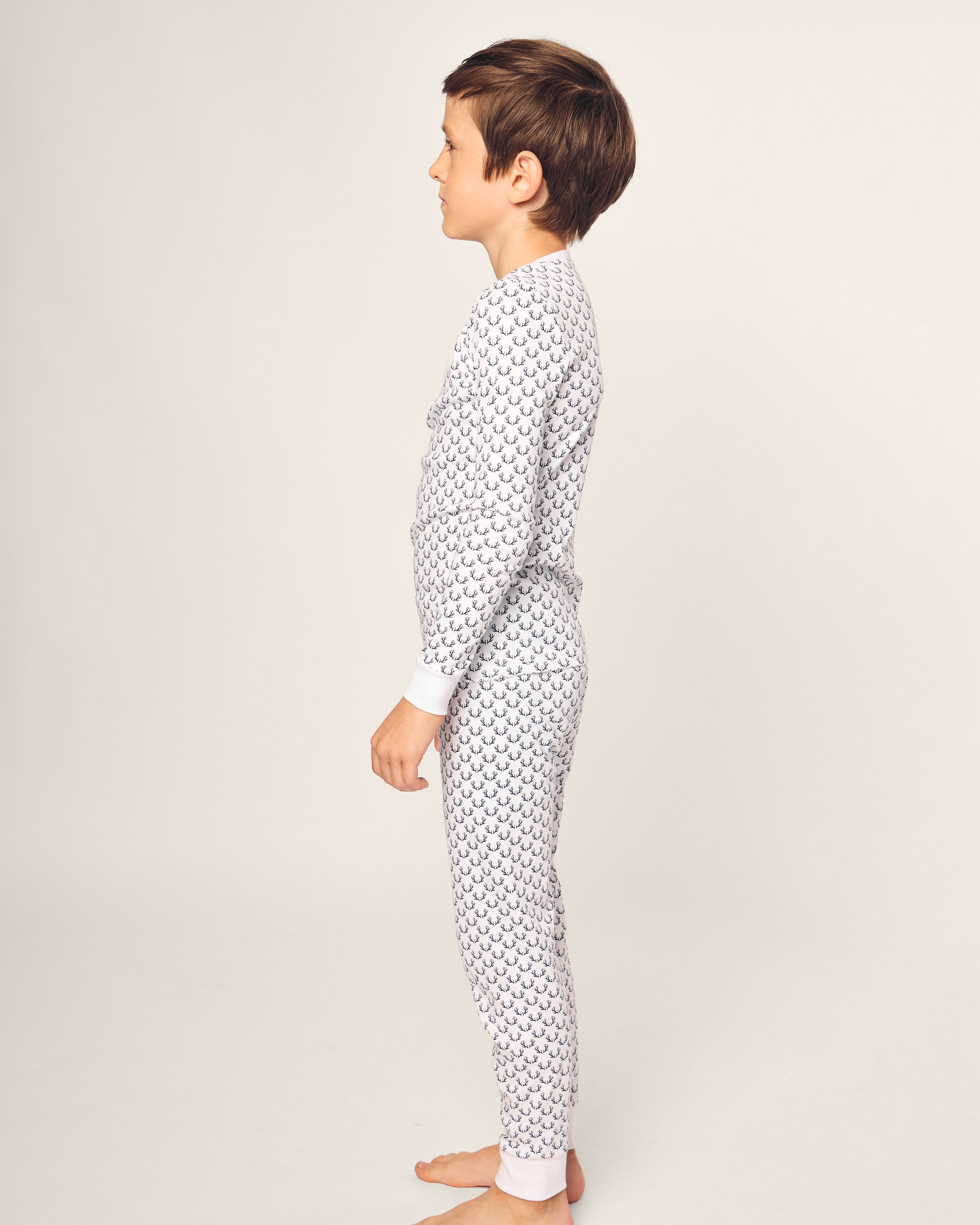 A child in profile on a plain background wears the Petite Plume Kids Pima Snug Fit Pajama Set in Nordic Antlers on White. The soft Pima Cotton sleepwear covers the whole body with a subtle pattern. The child is barefoot and looking forward.