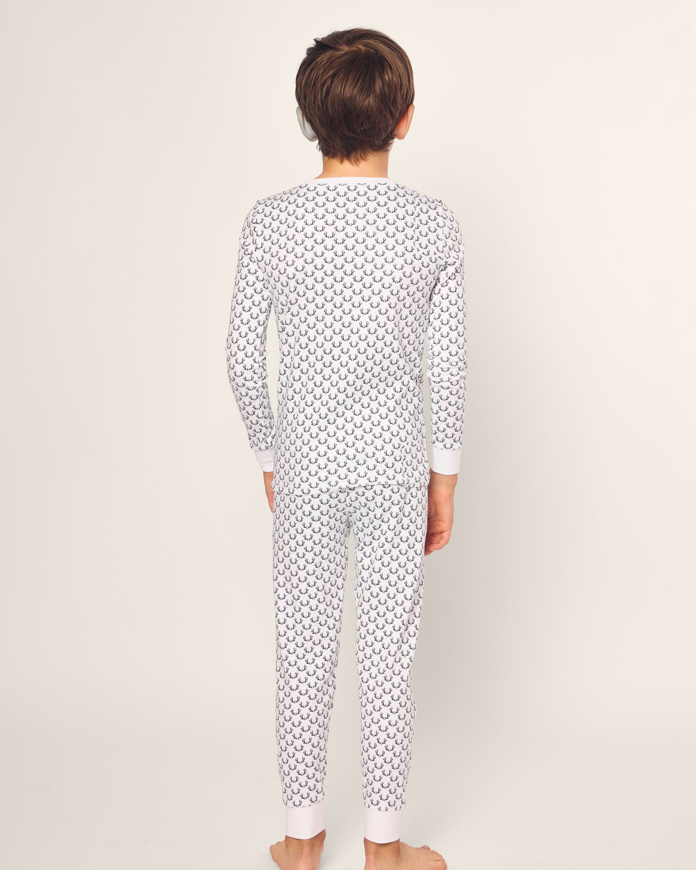 A child with short brown hair stands barefoot, facing away from the camera in a Petite Plume white Pima Snug Fit Pajama Set, featuring the Nordic Antlers design. The background is plain and light-colored.