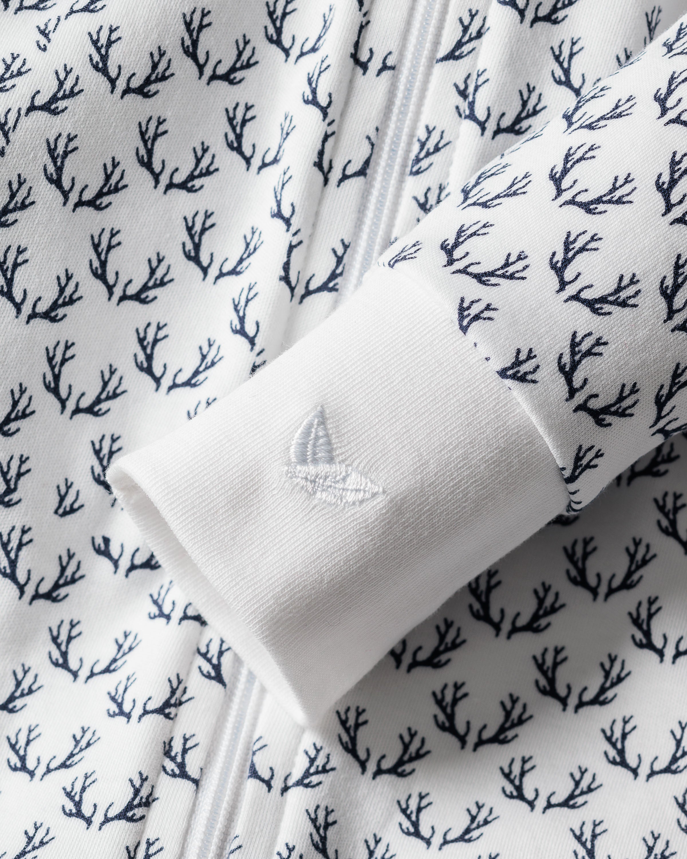 Close-up of Petite Plumes Babys Pima Romper in Nordic Antlers on White. The focus is on a romper sleeve with an embroidered sailboat near the cuff, perfect for elegant sleepwear enthusiasts.