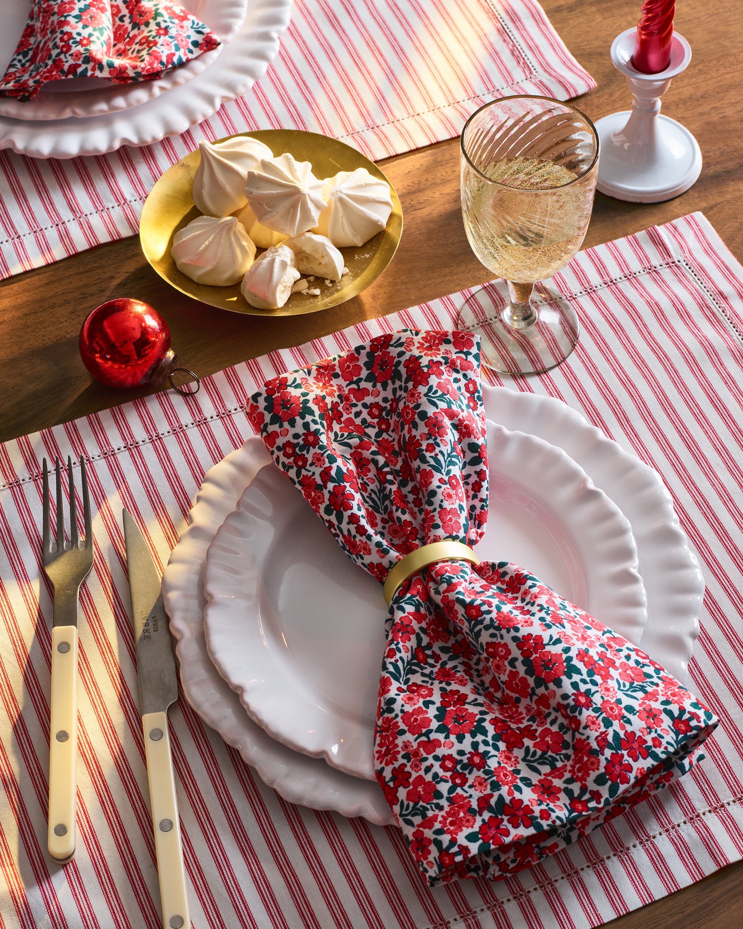 The table setting includes a white plate and red floral napkin, elevated by elegant linens and cream-handled utensils. Featuring Petite Plumes Signature Twill Placemats in Antique Red Ticking, it is adorned with white wine, meringues, a red ornament, and candle on the striped cloth.
