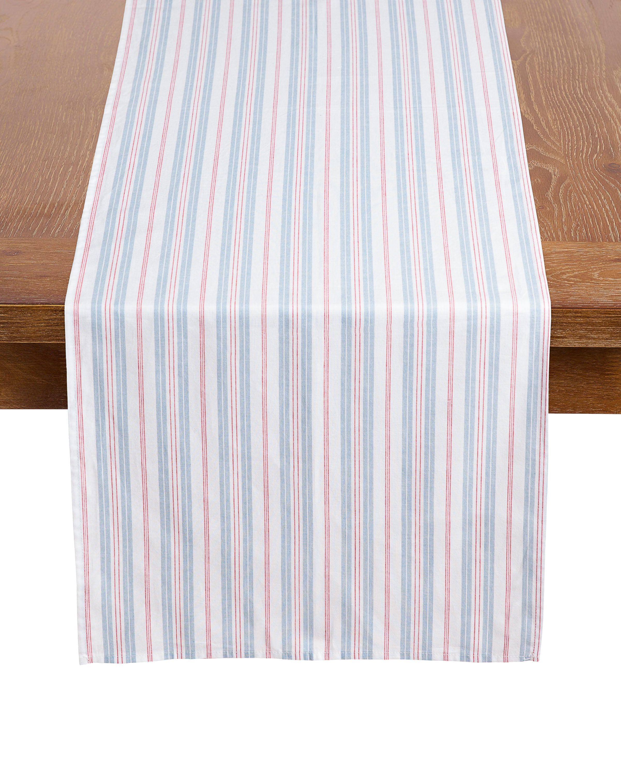A folded and an unfolded Signature Twill Table Runner & Napkin set in Vintage French Stripes, featuring blue, red, and white vertical stripes, displayed on a wooden table and against a white background.