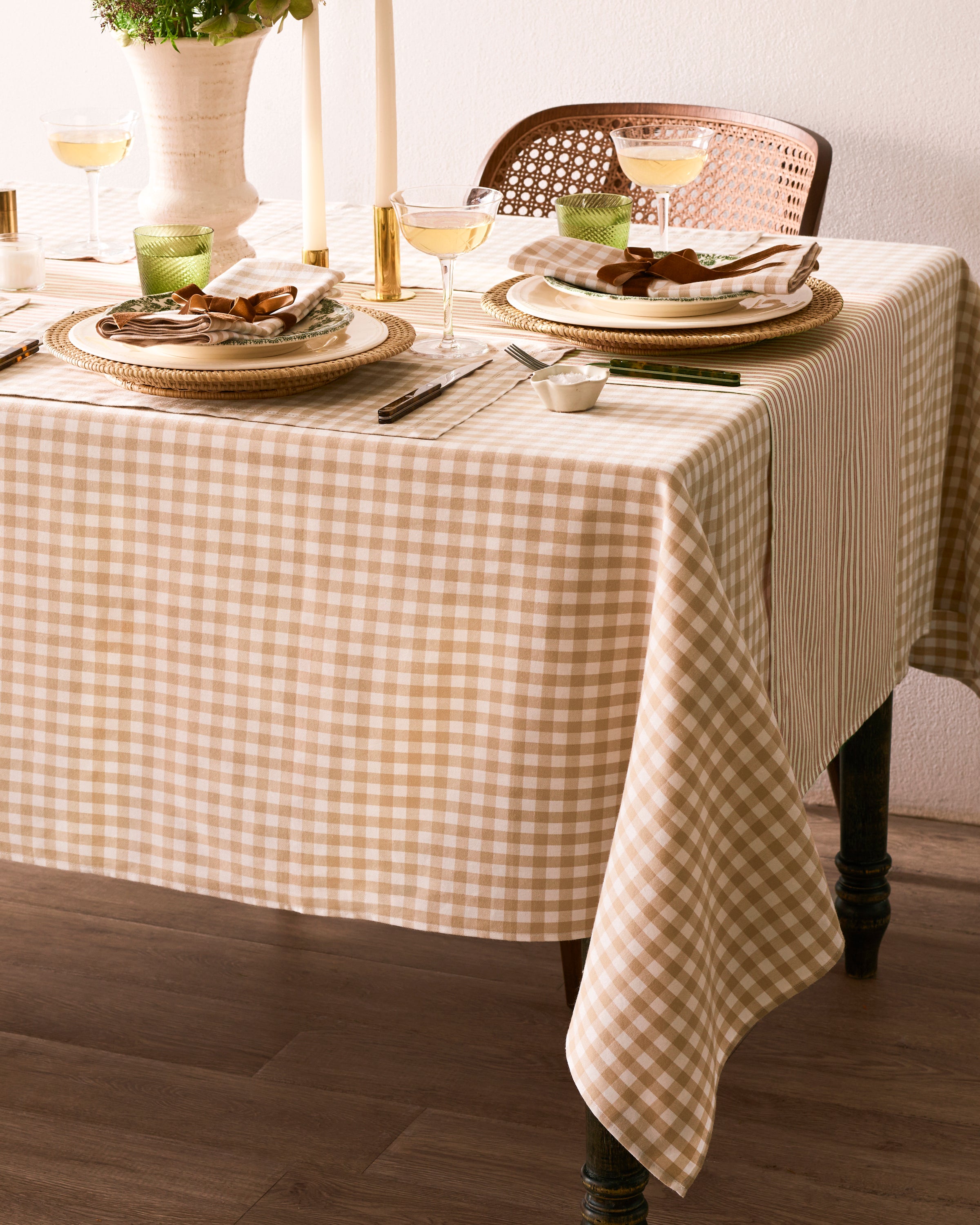 The Signature Twill Tablecloth in Camel Gingham, featuring beige and white checks, covers a rectangular table with wooden legs and drapes over the sides. The background is plain white.
