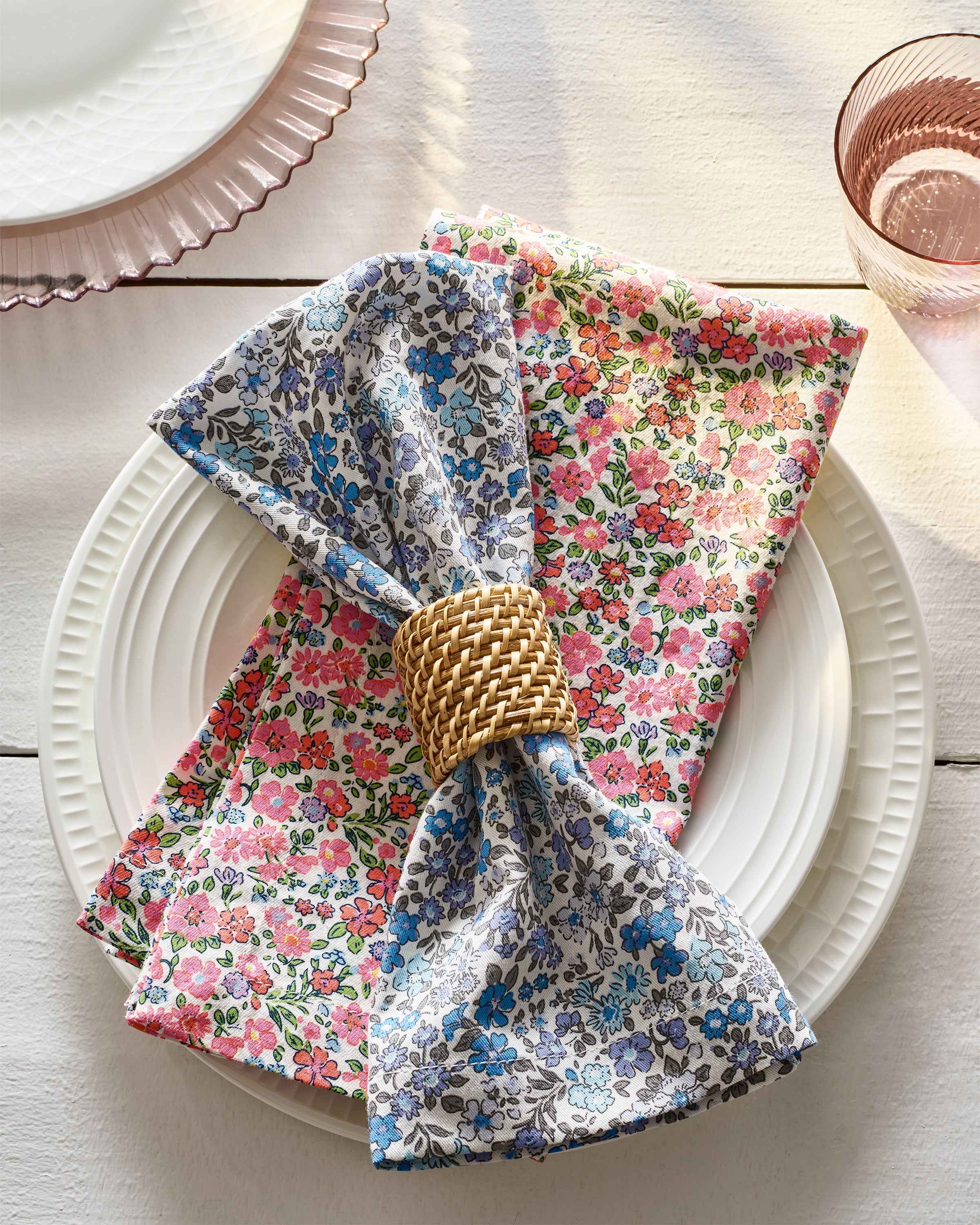 A white plate rests on a light wooden table with a Petite Plume Signature Twill Napkin in Fleur DAzur, held by a woven ring. Made from 100% cotton, the napkin features charming prints. A glass with a beverage is partially visible beside it.