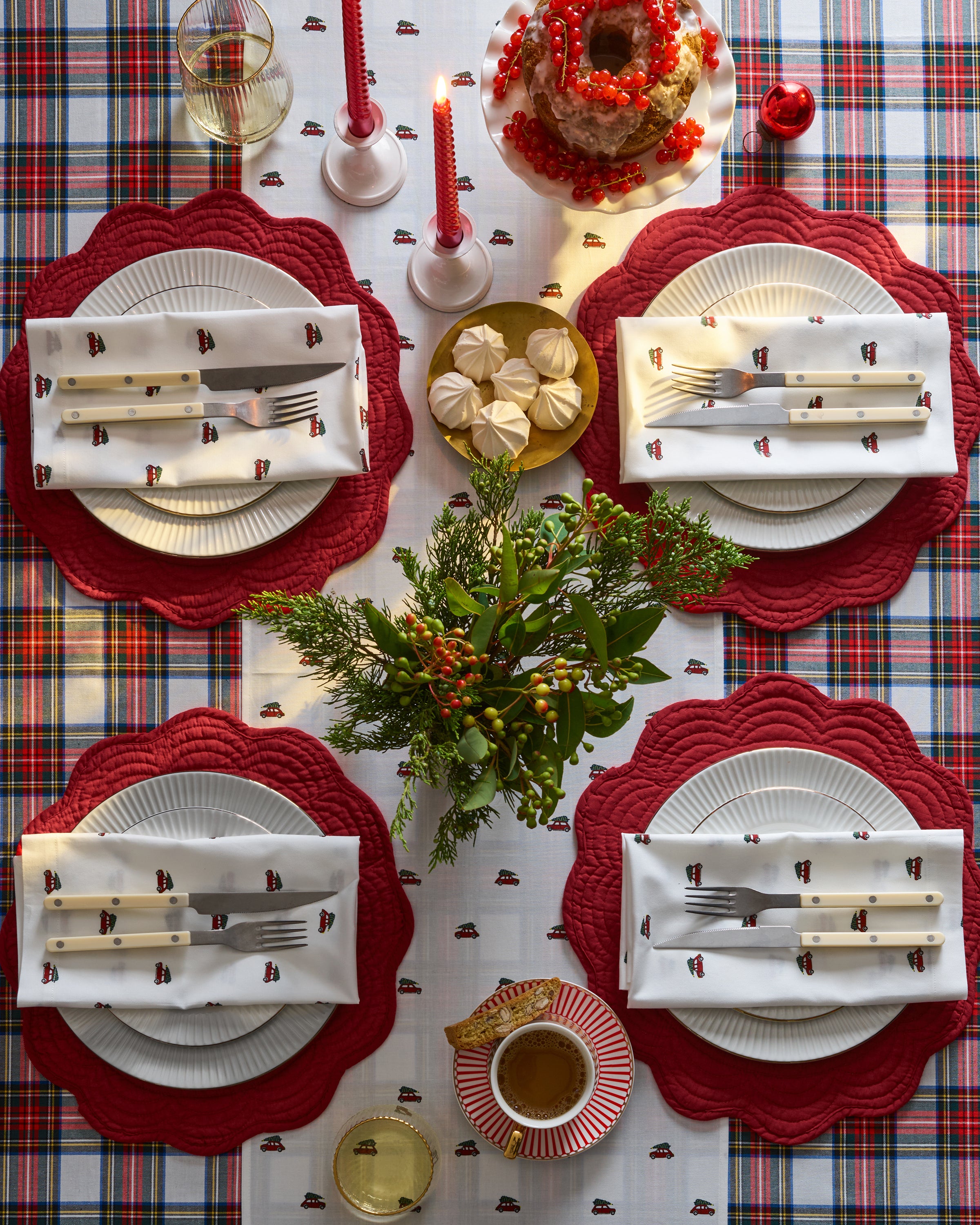 A festive tablescape with a red, white, and plaid theme showcases Petite Plumes 100% cotton Signature Twill Table Runner in Holiday Journey. Four place settings include Christmas-themed napkins, red placemats, and silverware. The centerpiece features greenery, two red candles, and a nearby dessert.