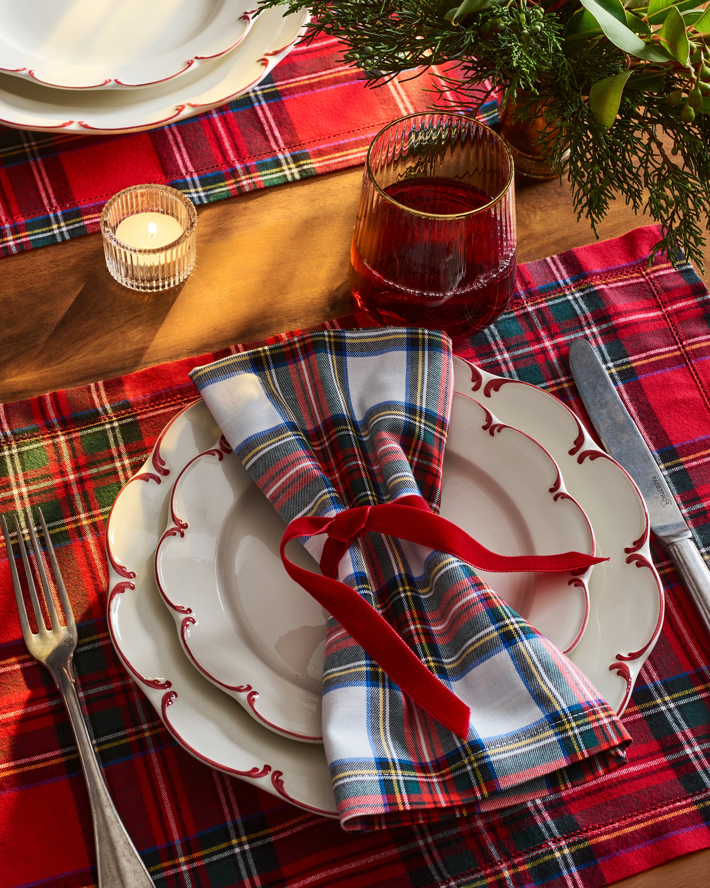 A festive table setting features Petite Plumes Signature Twill Placemats in Imperial Tartan, white scalloped plates, a red ribbon, and a glass of red drink. A small candle and greenery complete the warm, holiday-themed decor with versatile prints perfect for any celebration.