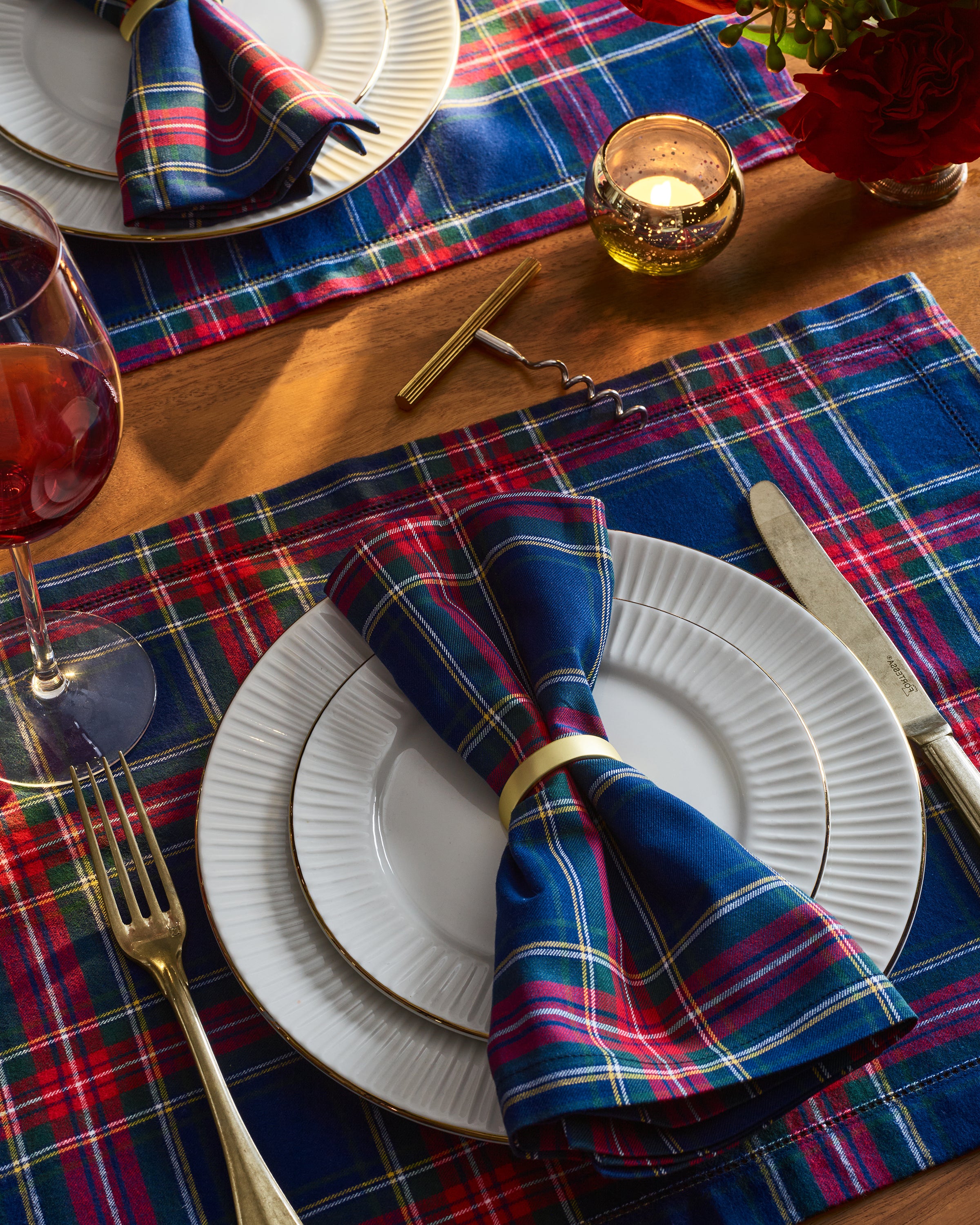 A cozy tablescape features white plates on Signature Twill Placemats in Windsor Tartan by Petite Plume, each with a matching napkin tied in a gold ring. A wine glass, fork, knife, corkscrew, candle, and vibrant bouquet of red flowers complete the look with versatile red, blue, and green hues.