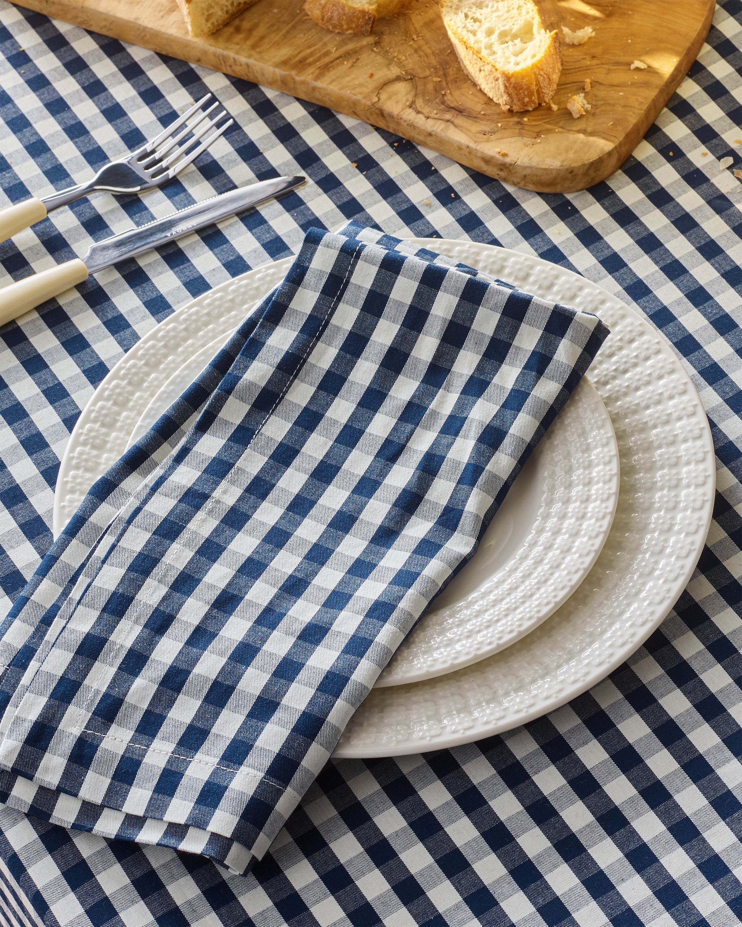 A table is adorned with Petite Plumes Signature Twill Napkins in Navy Gingham. A white textured dinner plate is stacked on a smaller one, silverware alongside. A wooden board holds sliced bread in the background.