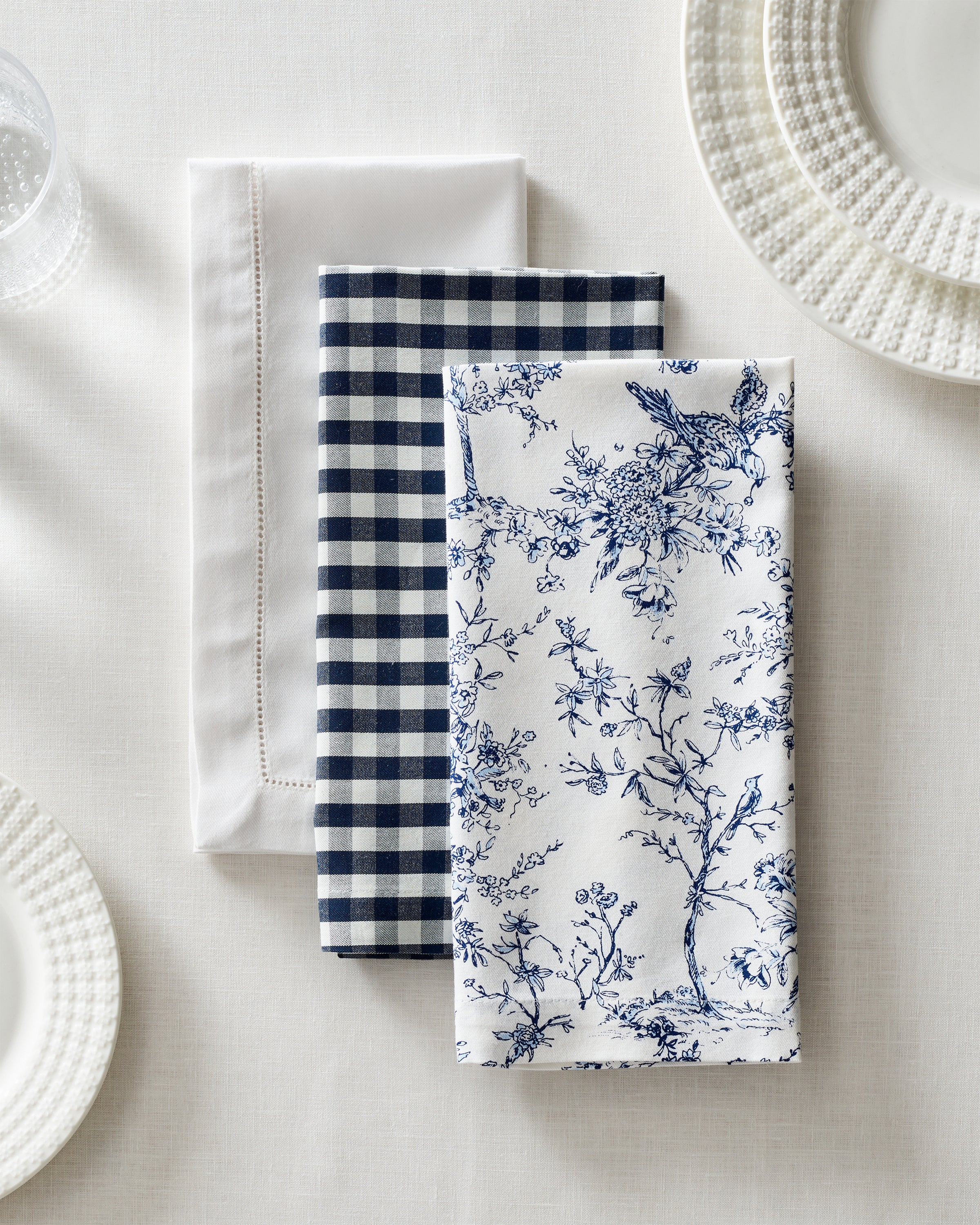 Three Signature Twill Napkins by Petite Plume, crafted from 100% cotton, sit on a white tablecloth. Each has a unique design: solid white, navy gingham, and blue floral. Two textured white plates peek out, enhancing the elegant display of table linens.