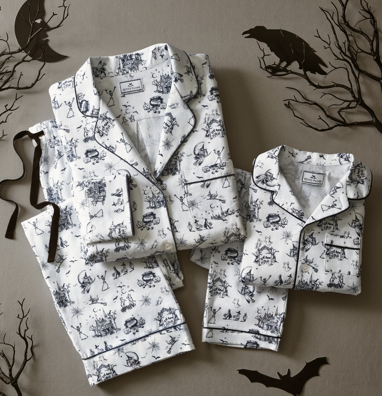 Matching adult and child pajamas with black Halloween-themed prints, laid on a gray surface decorated with cutout bats, trees, and ravens for a spooky atmosphere.