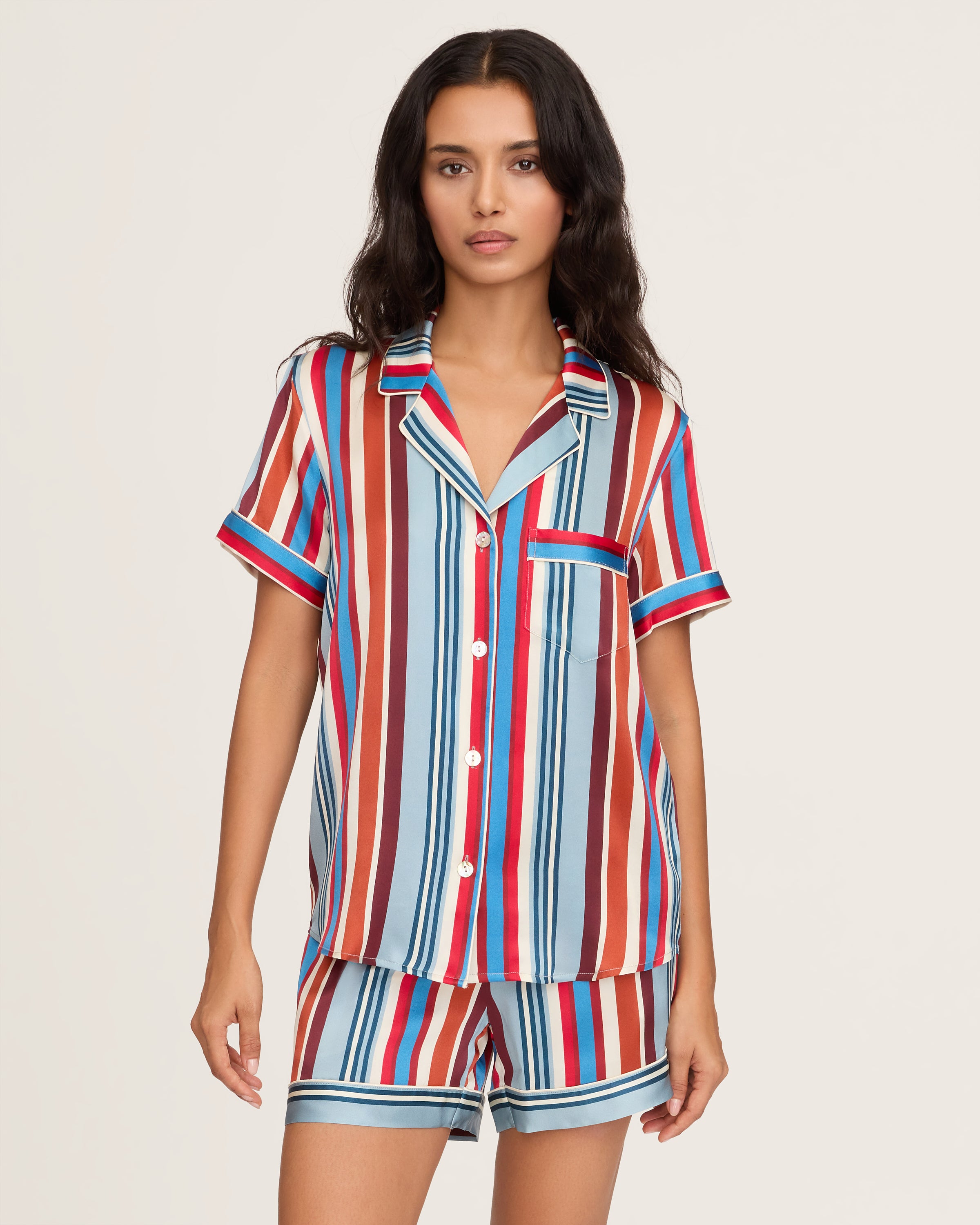 The Women's Silk Short Set in Discovery Stripe features a short-sleeved button-up shirt and matching drawstring shorts with bold vertical stripes in red, blue, brown, and white.
