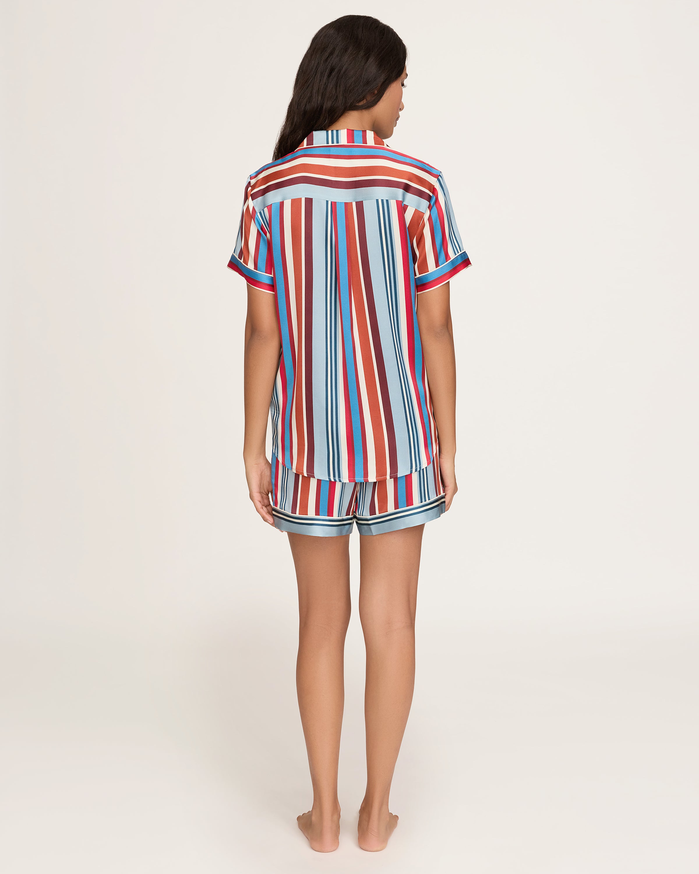The Women's Silk Short Set in Discovery Stripe features a short-sleeved button-up shirt and matching drawstring shorts with bold vertical stripes in red, blue, brown, and white.