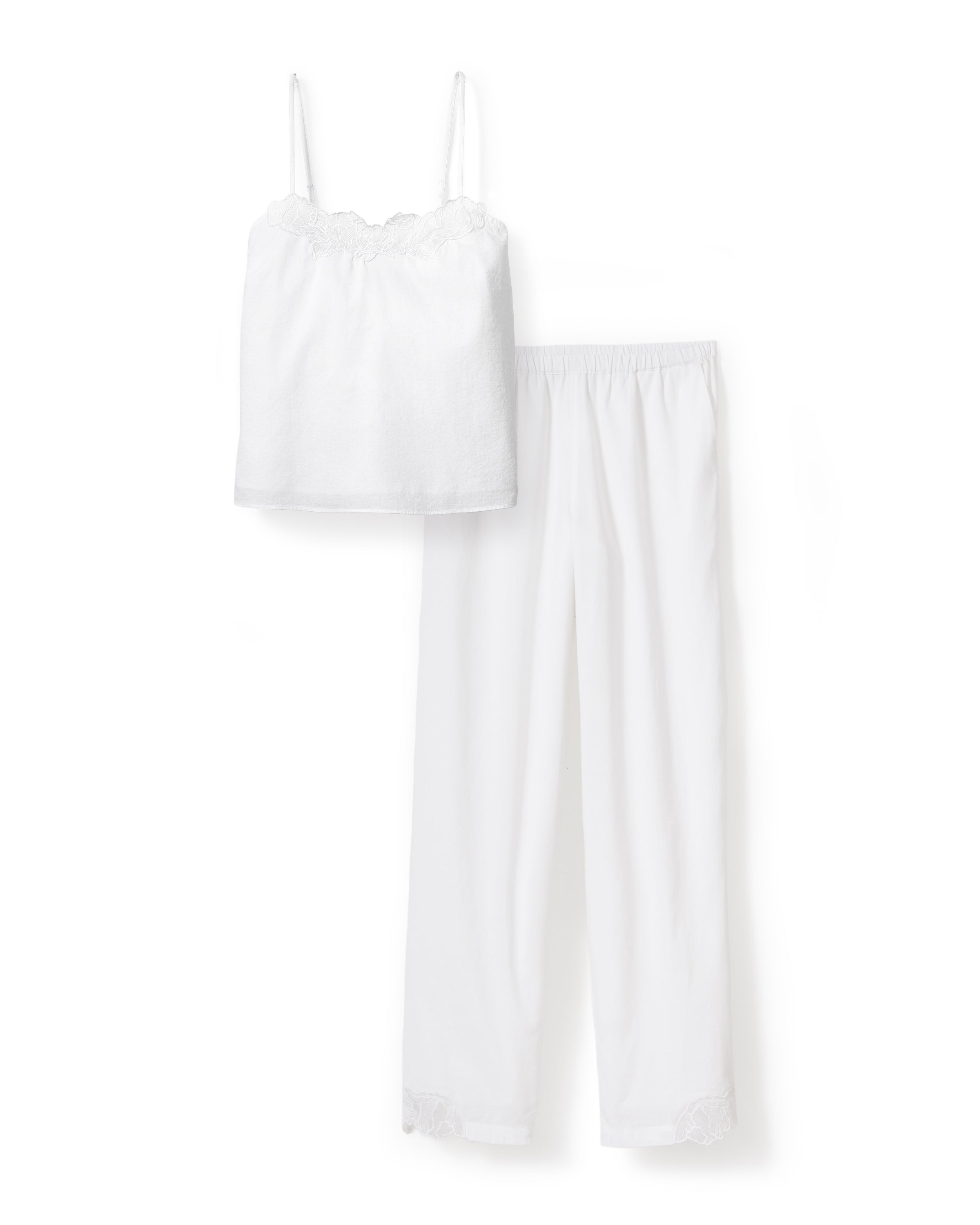 A woman with wavy hair stands barefoot on a light background, wearing the Women's Voile Fleur Cami Pant Set in White—featuring a sleeveless top and pants with lace trim. Her arms are behind her back.