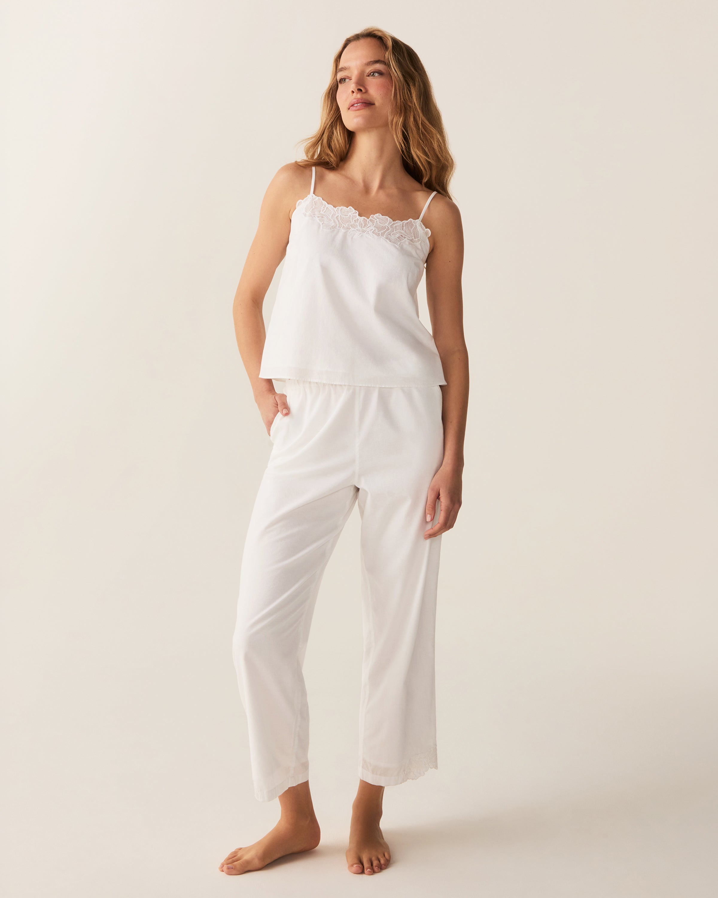 A woman with wavy hair stands barefoot on a light background, wearing the Women's Voile Fleur Cami Pant Set in White—featuring a sleeveless top and pants with lace trim. Her arms are behind her back.