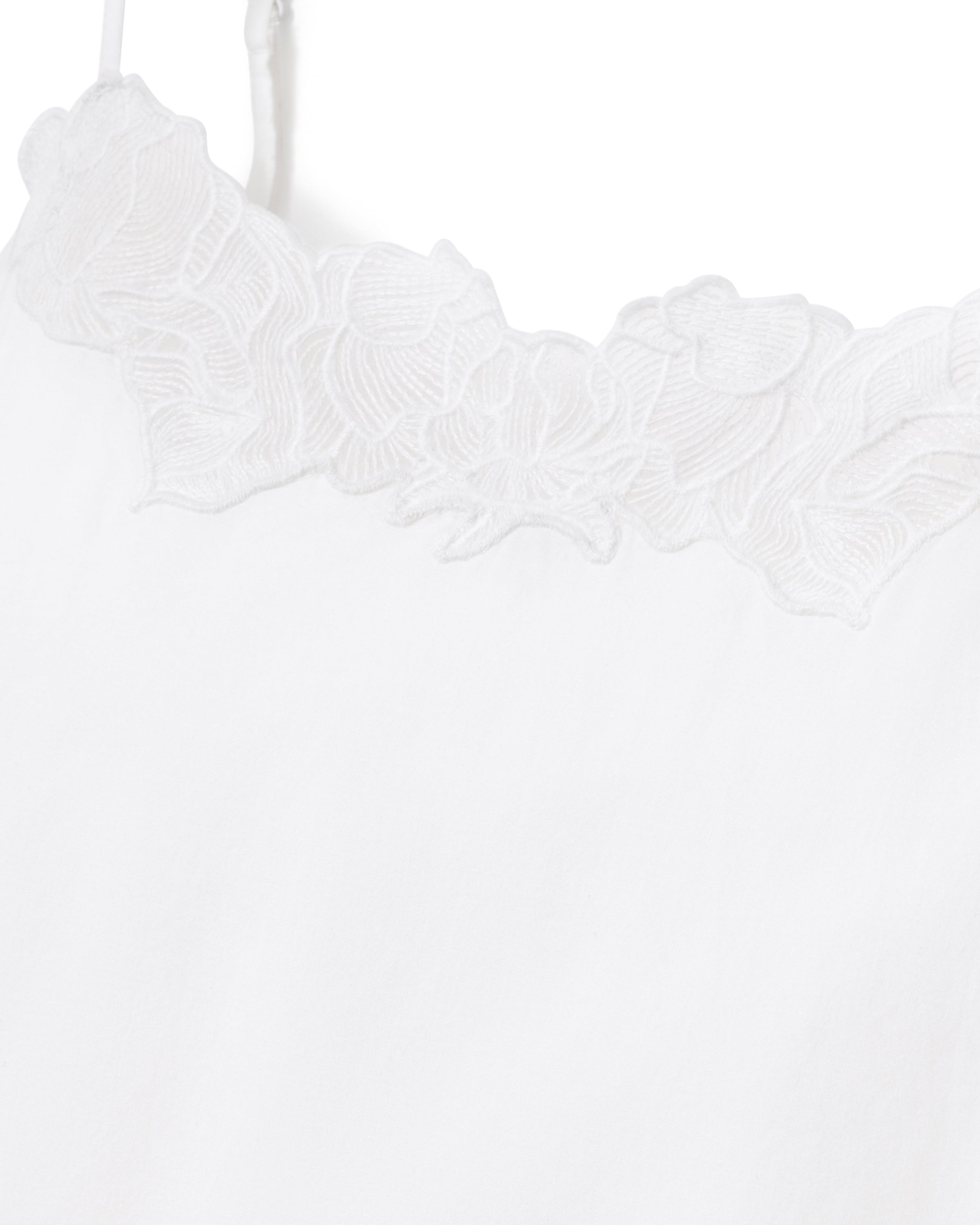 A woman with wavy hair stands barefoot on a light background, wearing the Women's Voile Fleur Cami Pant Set in White—featuring a sleeveless top and pants with lace trim. Her arms are behind her back.