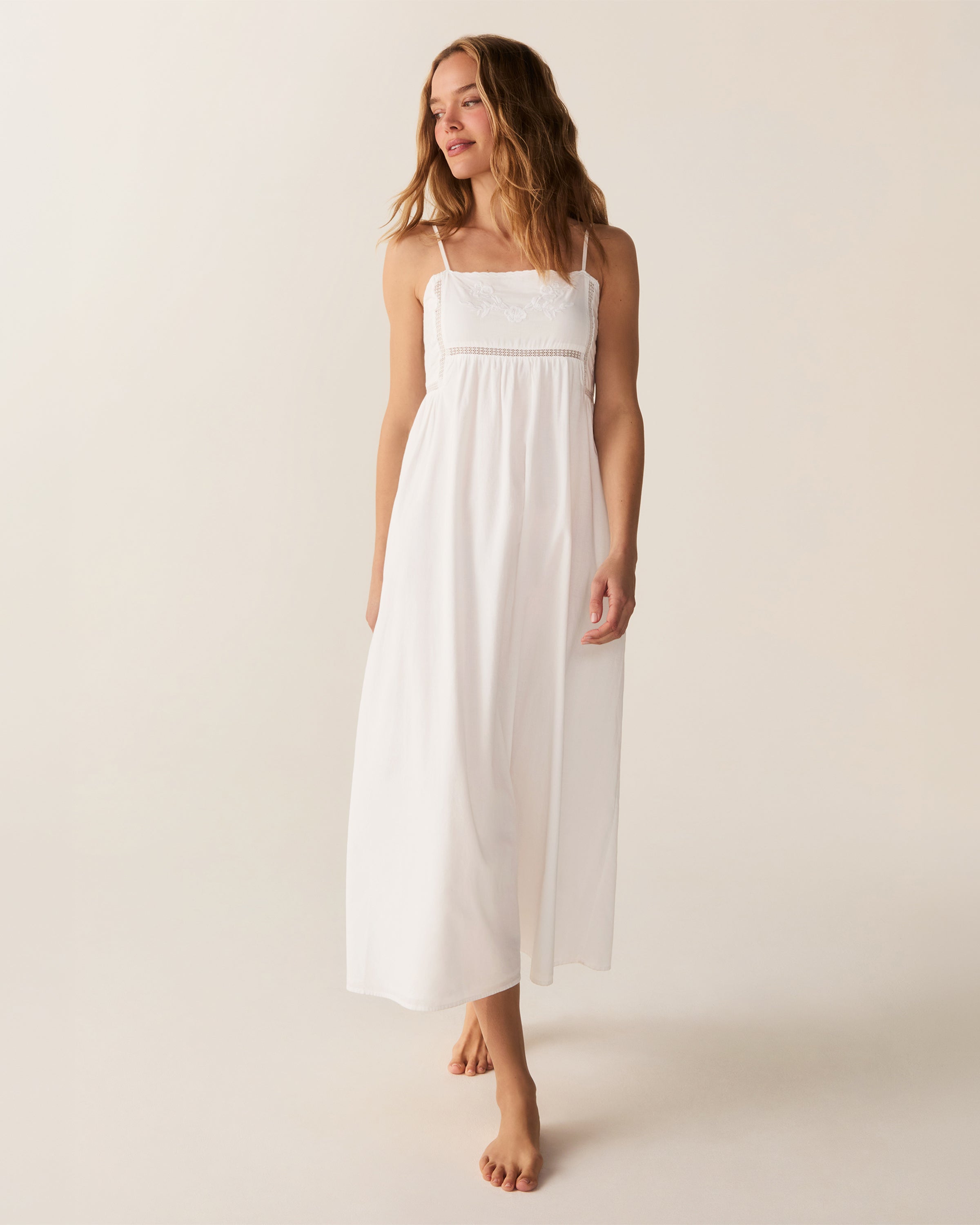 A woman with wavy hair stands barefoot and smiling to the side, wearing the Women's Voile Luna Gown in White—a long, sleeveless dress with thin straps—against a plain, light-colored background.