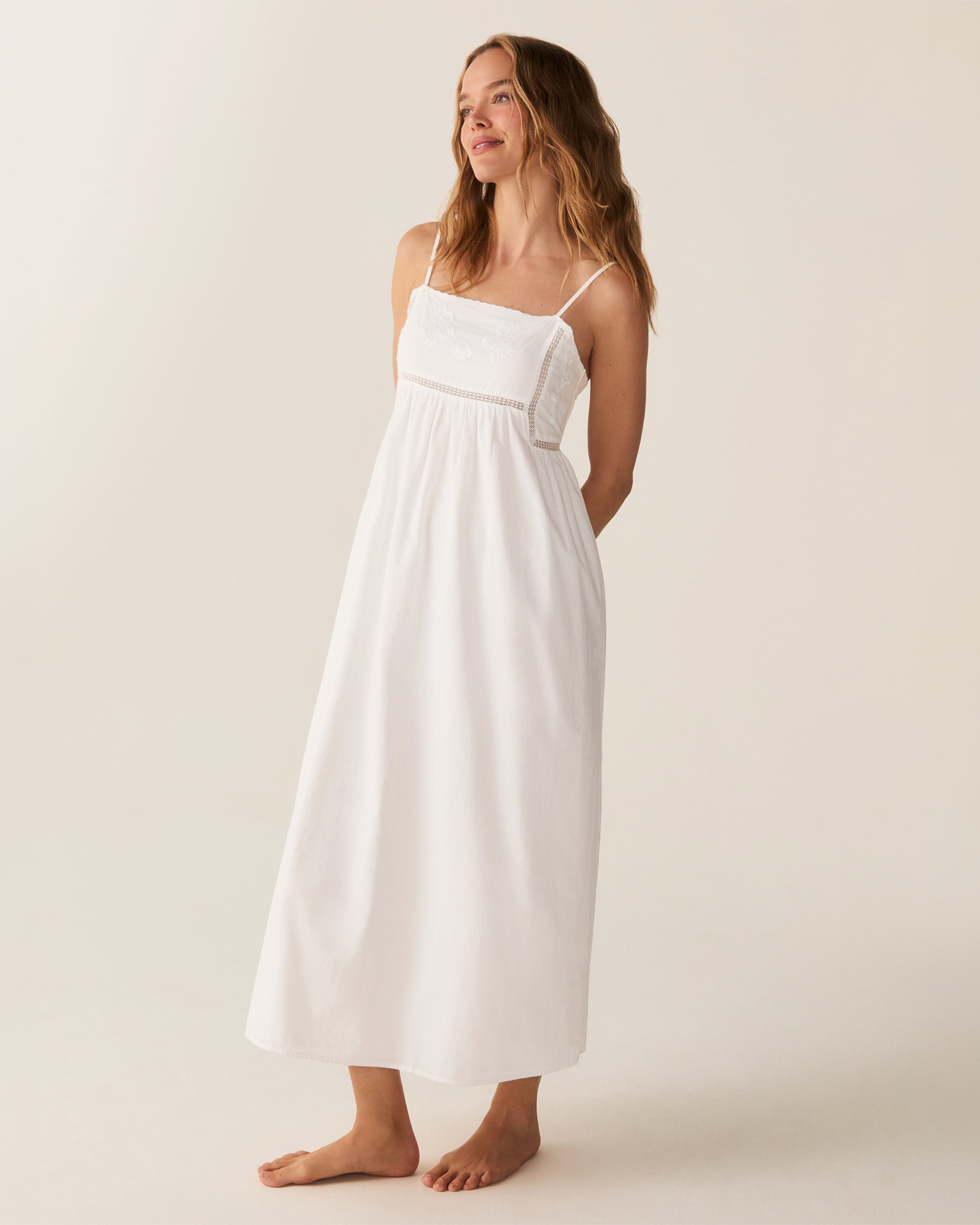 A woman with wavy hair stands barefoot and smiling to the side, wearing the Women's Voile Luna Gown in White—a long, sleeveless dress with thin straps—against a plain, light-colored background.