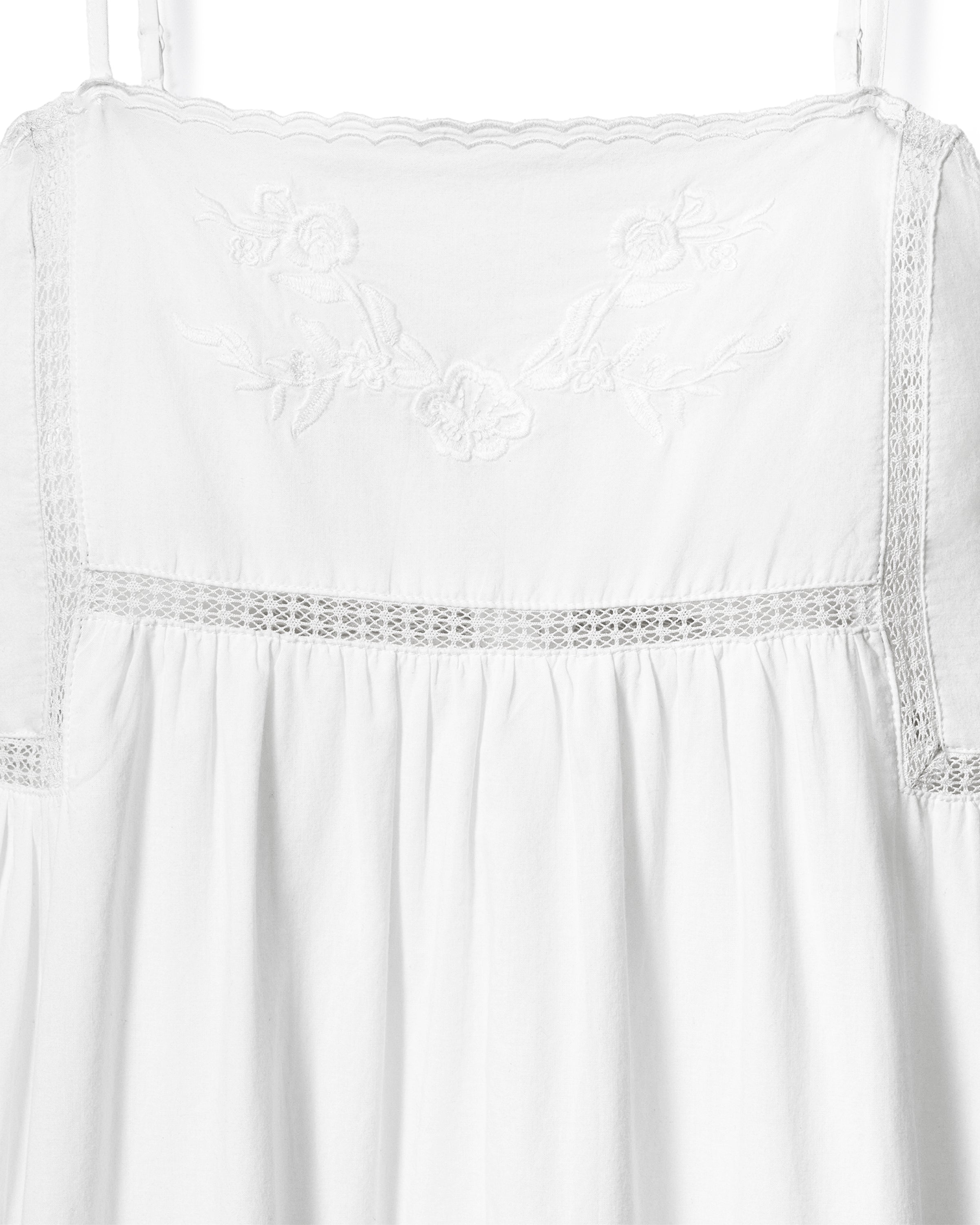 A woman with wavy hair stands barefoot and smiling to the side, wearing the Women's Voile Luna Gown in White—a long, sleeveless dress with thin straps—against a plain, light-colored background.