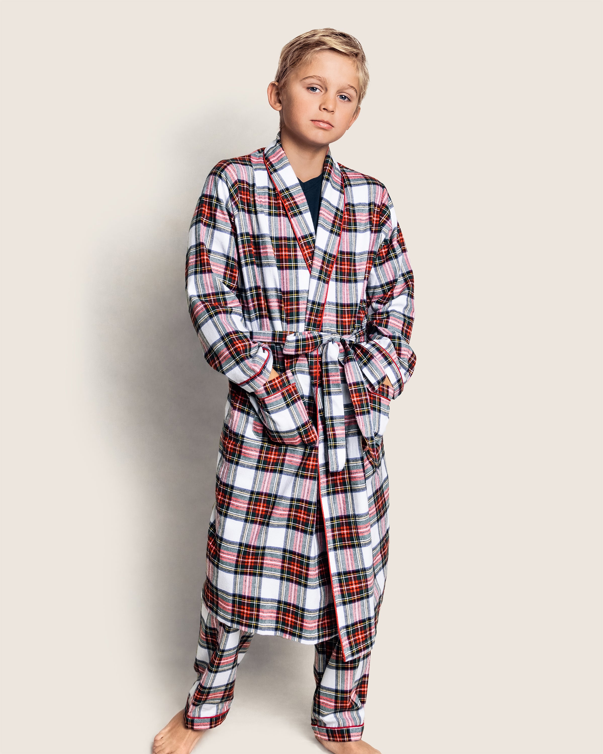 A child in Petite Plumes Kids Brushed Cotton Robe in Balmoral Tartan stands barefoot against a plain backdrop, hands in pockets. The cozy plaid robe and matching pants, tied with a belt, feature a rich red, white, and black design—ideal for comfy nights.