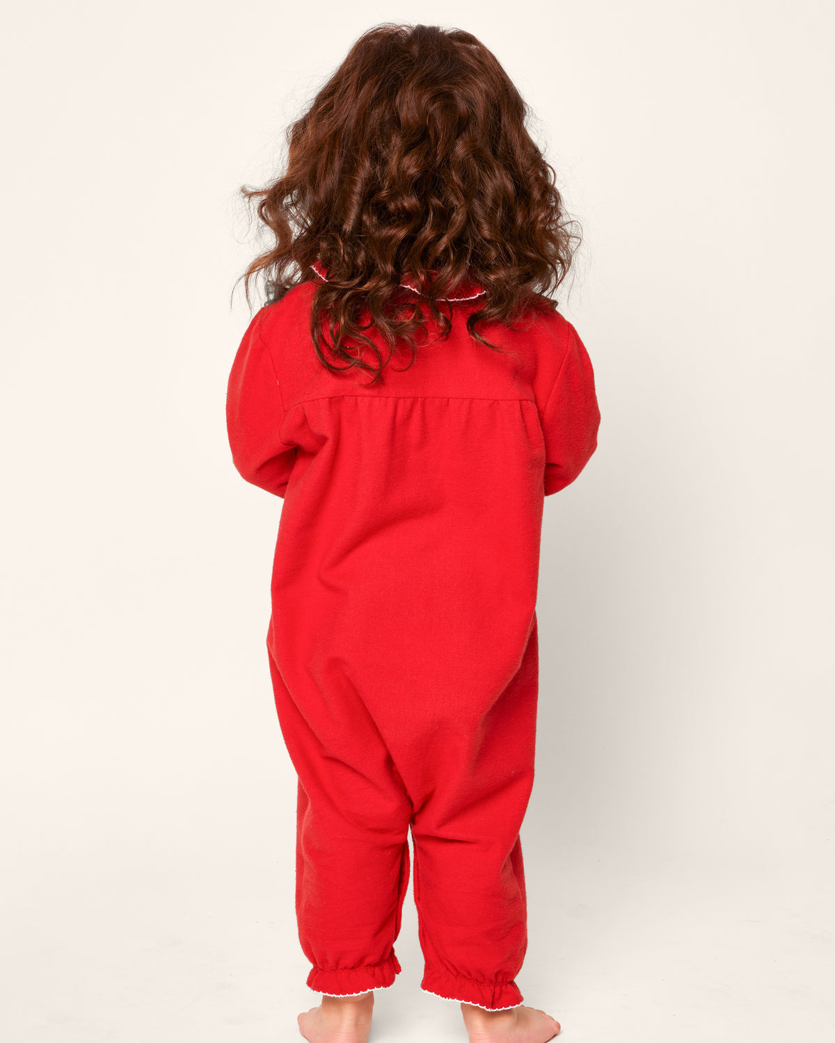 A child stands barefoot against a plain off-white background, wearing the Babys Flannel Madeline Romper in Red by Petite Plume. The romper is made from flame-retardant fabric. Curly brown hair cascades down their back.