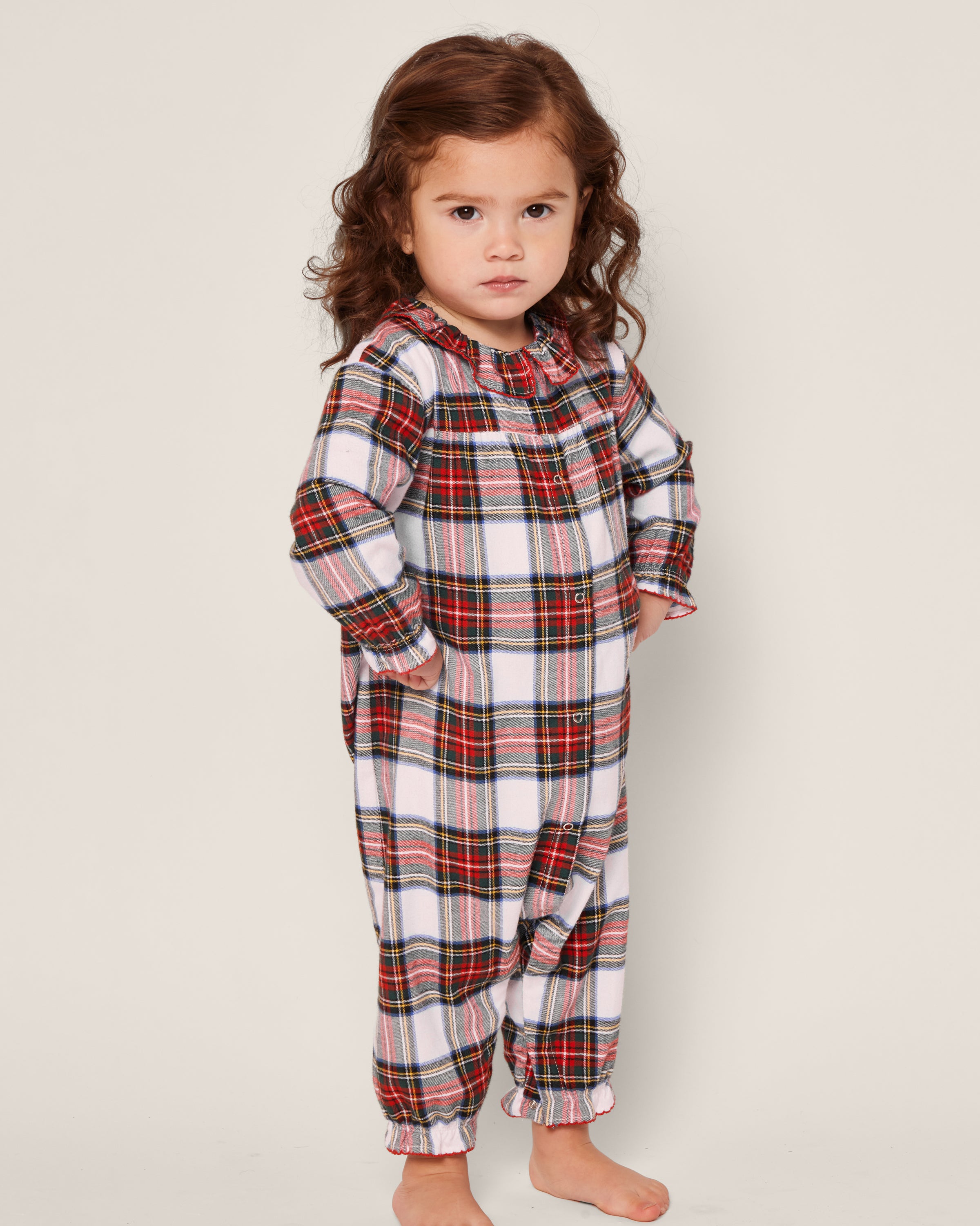 A young child with curly brown hair, hands on hips, seriously poses barefoot against a plain background, wearing Petite Plumes Babys Brushed Cotton Madeline Romper in a cozy red, green, and white Balmoral Tartan pattern.