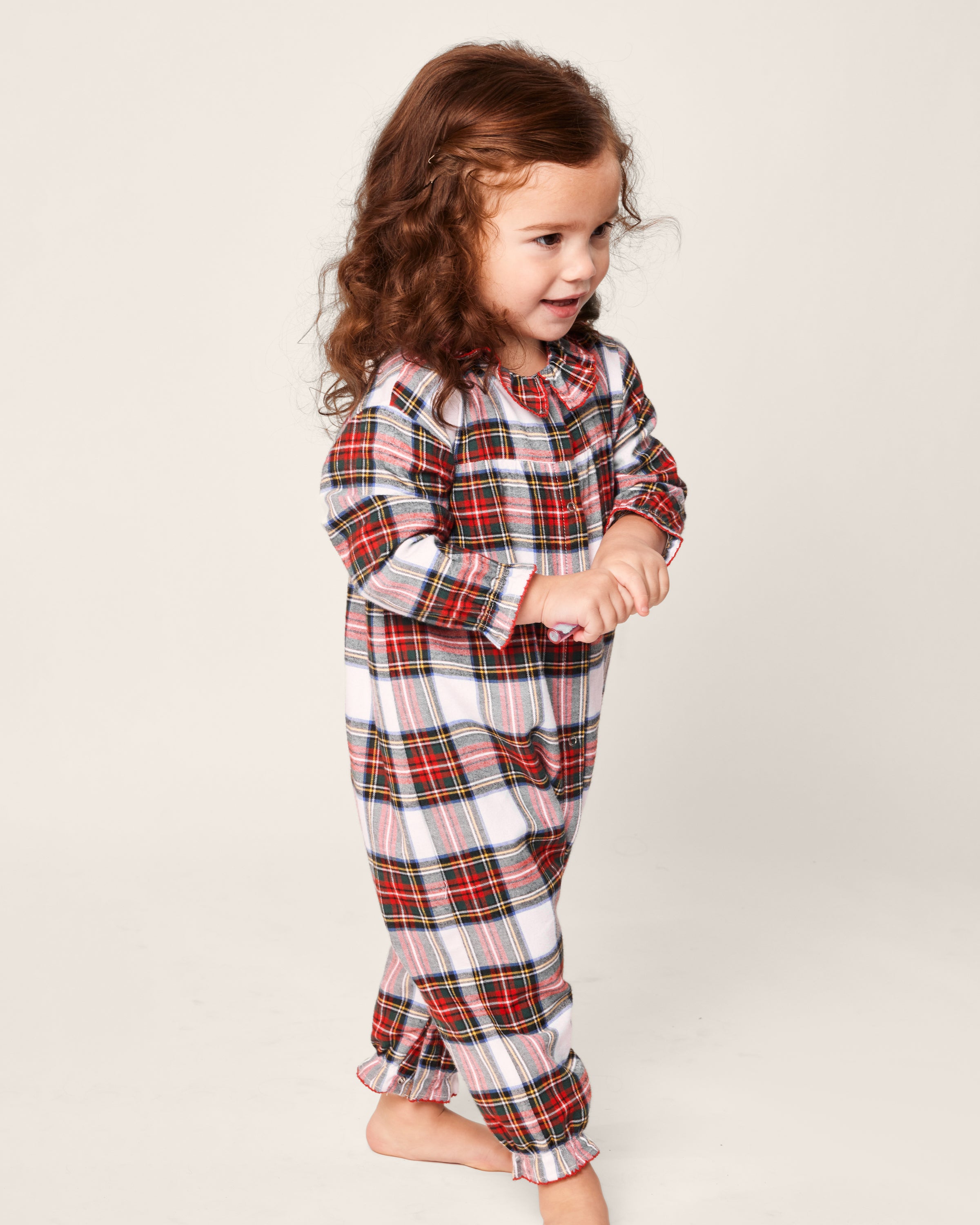 A young child with long brown hair beams in Petite Plumes Babys Brushed Cotton Madeline Romper, featuring flame-retardant Balmoral Tartan with red, green, and white plaid patterns. The little one smiles against a plain light-colored background.
