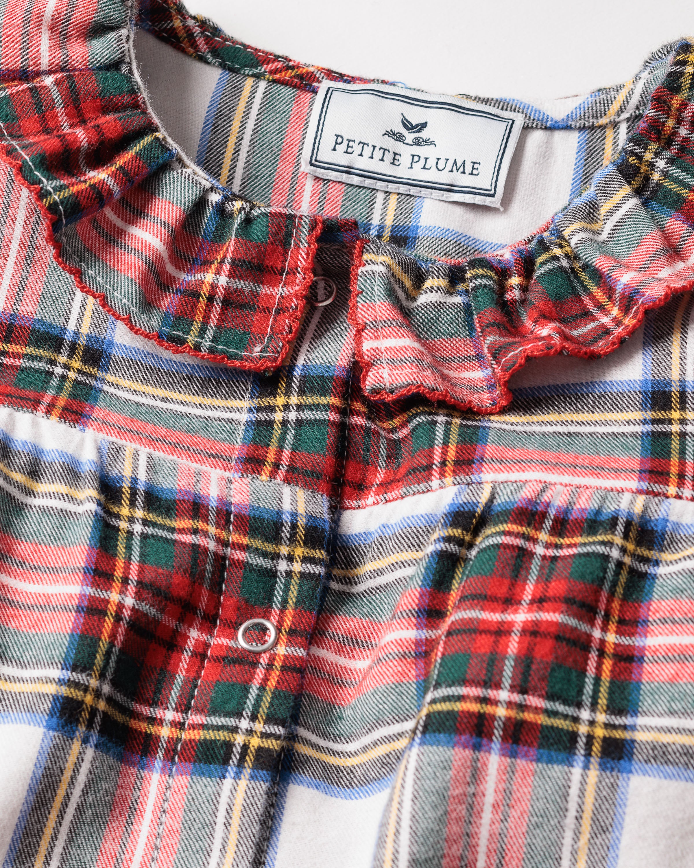 Close-up of the Babys Brushed Cotton Madeline Romper in Balmoral Tartan by Petite Plume. The plaid pattern showcases red, green, blue, and white hues. The romper is designed as childrens sleepwear with flame-retardant fabric and button closures. A bird graphic adorns the label inside the collar.