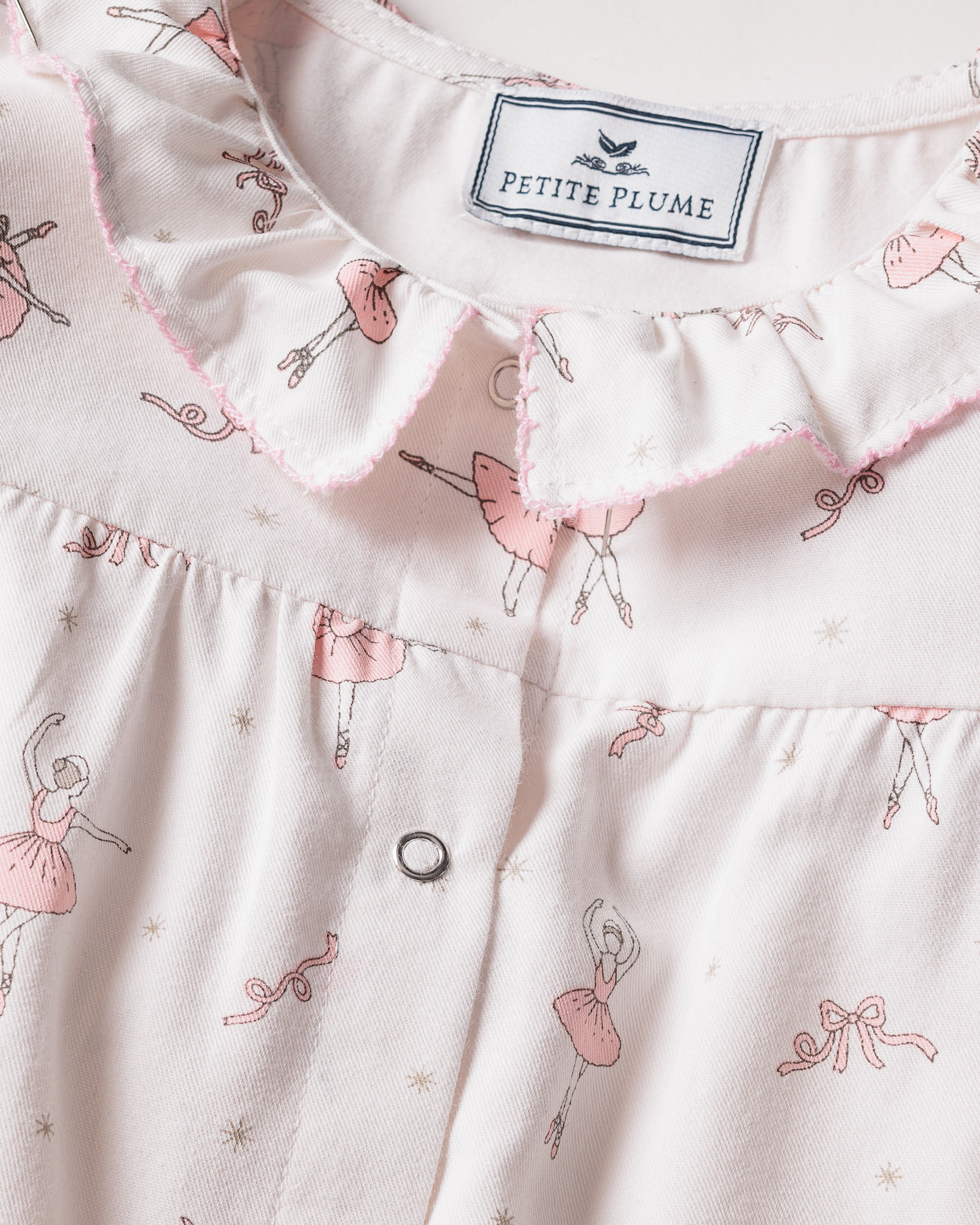 A close-up of Petite Plumes Babys Flannel Madeline Romper in Sugar Plum Fairy shows a light pink sleepwear garment with a frilled collar, ballet dancer print, flame-retardant fabric, delicate illustrations of Sugar Plum Fairies, small bows, and a single button.