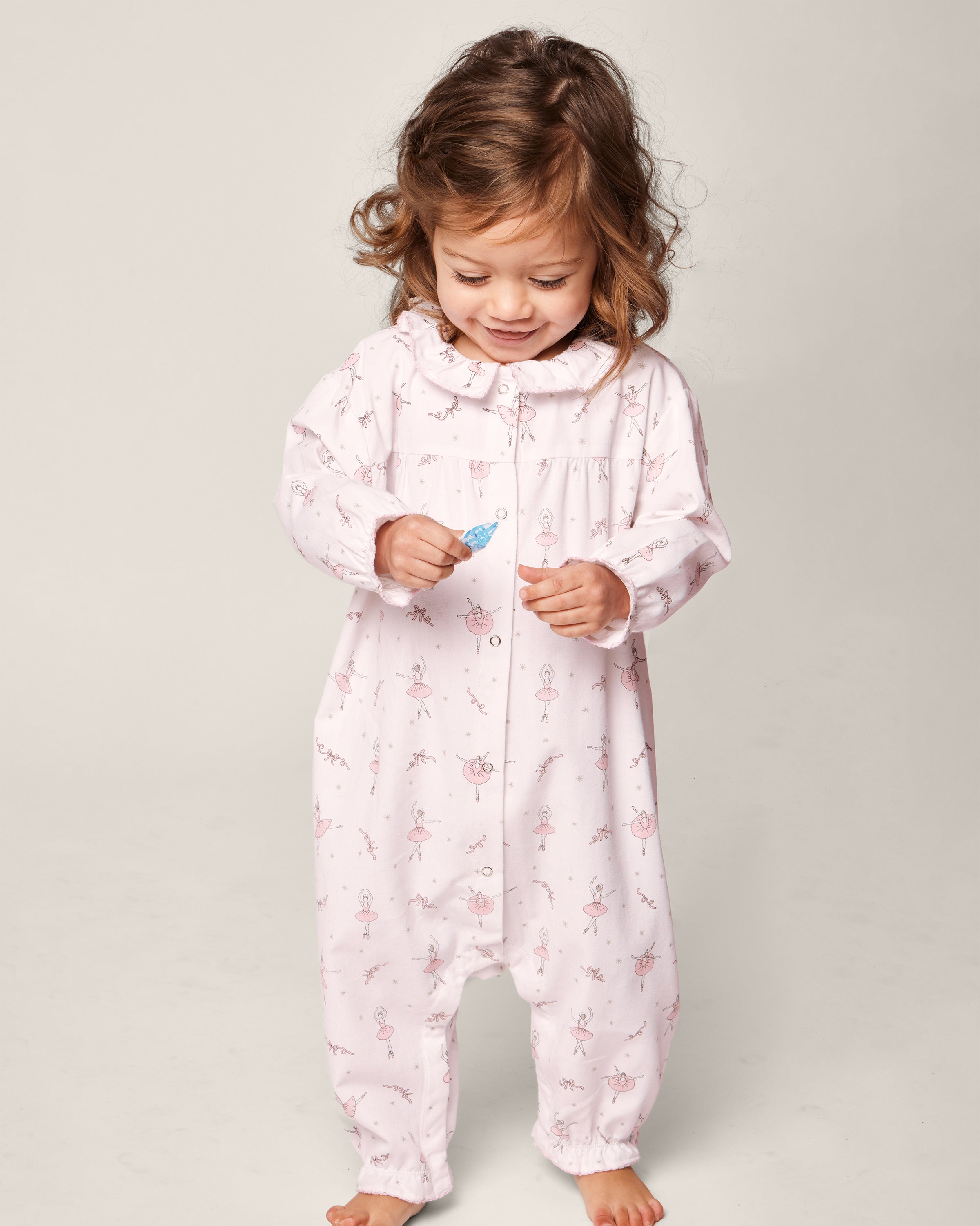 Standing barefoot against a neutral backdrop, a toddler wears Petite Plumes Babys Flannel Madeline Romper in Sugar Plum Fairy, crafted from flame-retardant fabric. They gaze down while holding a small toy.